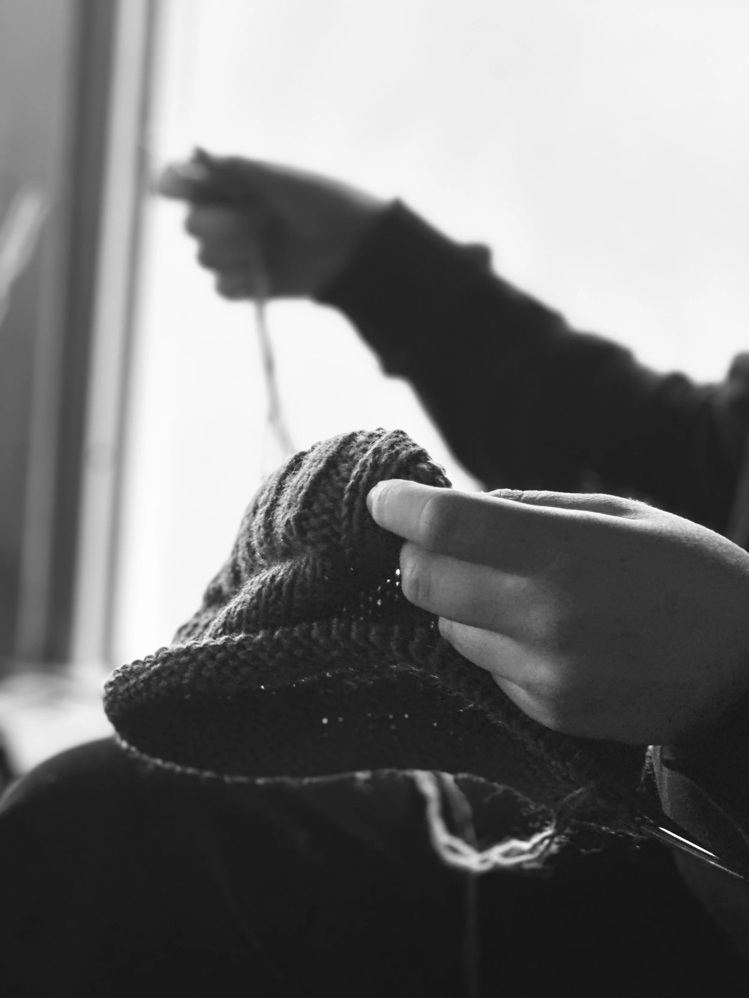 Close-up of a person knitting a hat with yarn, with another person's hand holding the hat, and a blurred background.