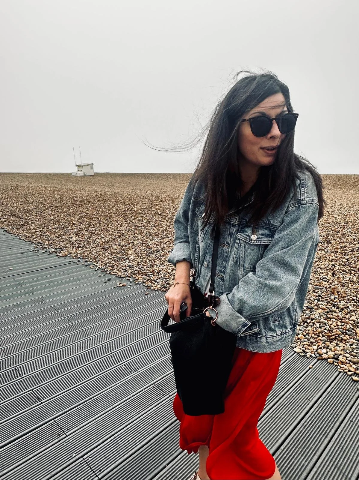 Woman with long dark hair wearing sunglasses, a denim jacket, and red pants, holding a black bag, standing on a boardwalk on a pebbled beach with overcast sky and a small white structure in the distance.