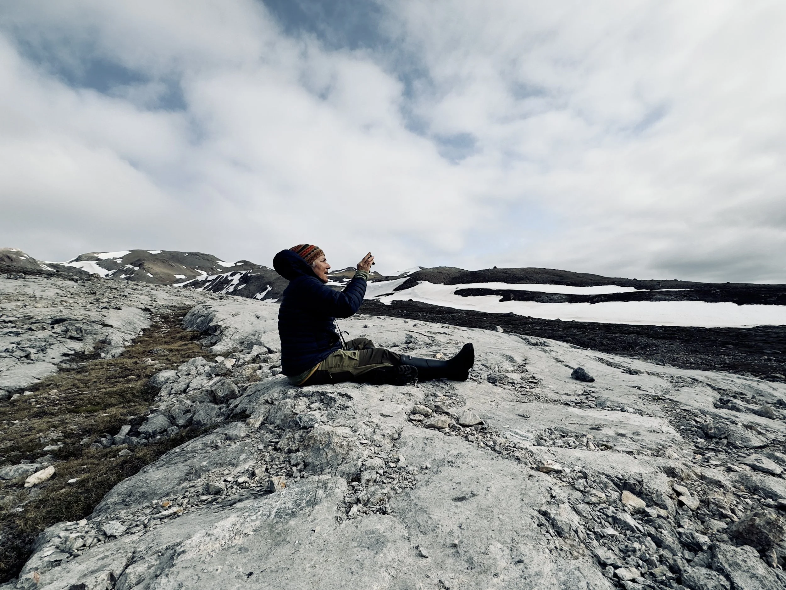 A woman sitting on rocky terrain in a mountainous landscape with patches of snow, taking a photo or video with her phone under a cloudy sky.