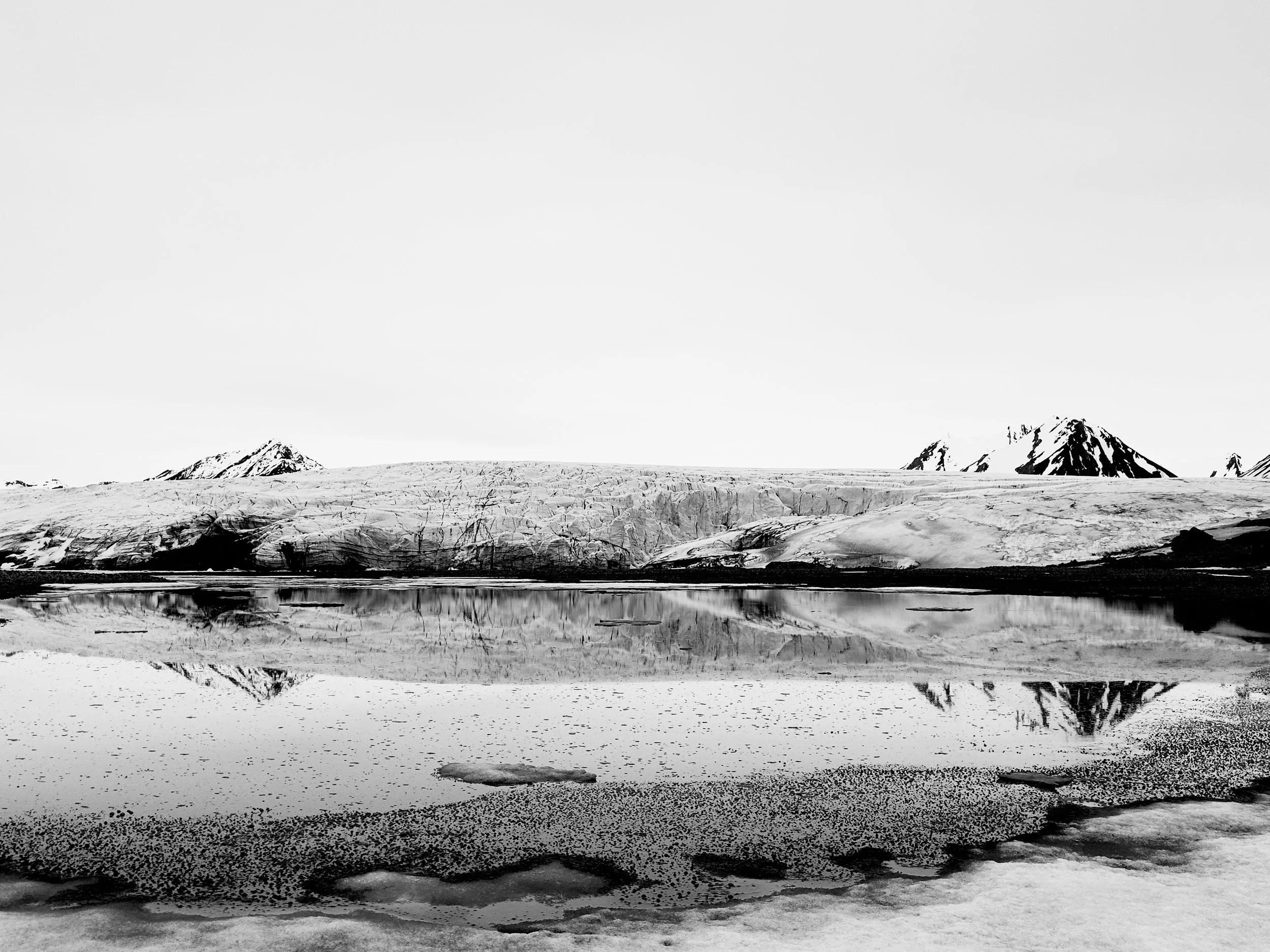 Black and white photo of a glacier and snow-covered mountains reflected in a body of water with ice on the shoreline.