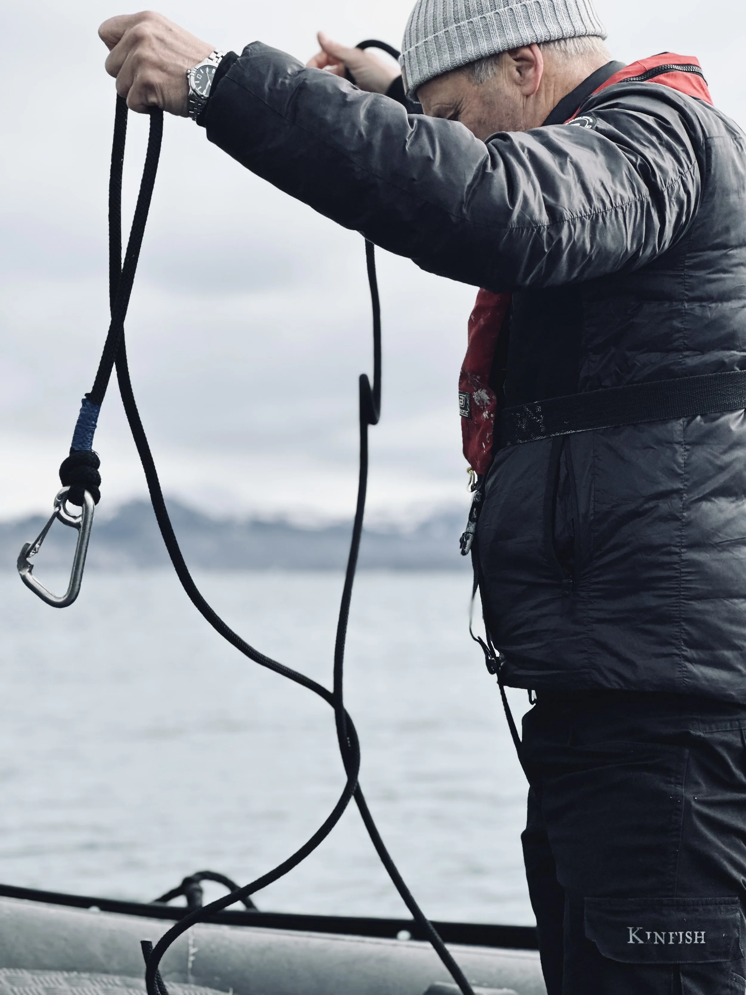 A man wearing a gray beanie, black jacket, and black pants with a 'KU FISH' label, holding a rope or cord with a carabiner near a body of water, with a distant shoreline and mountains in the background.