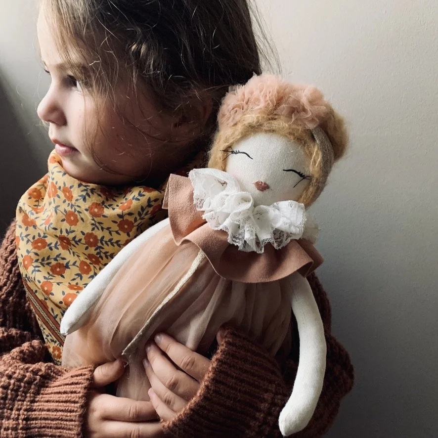 A young girl with brown hair wearing a floral orange and blue scarf holds a handmade cloth doll with pink hair, closed eyes, and a vintage dress.