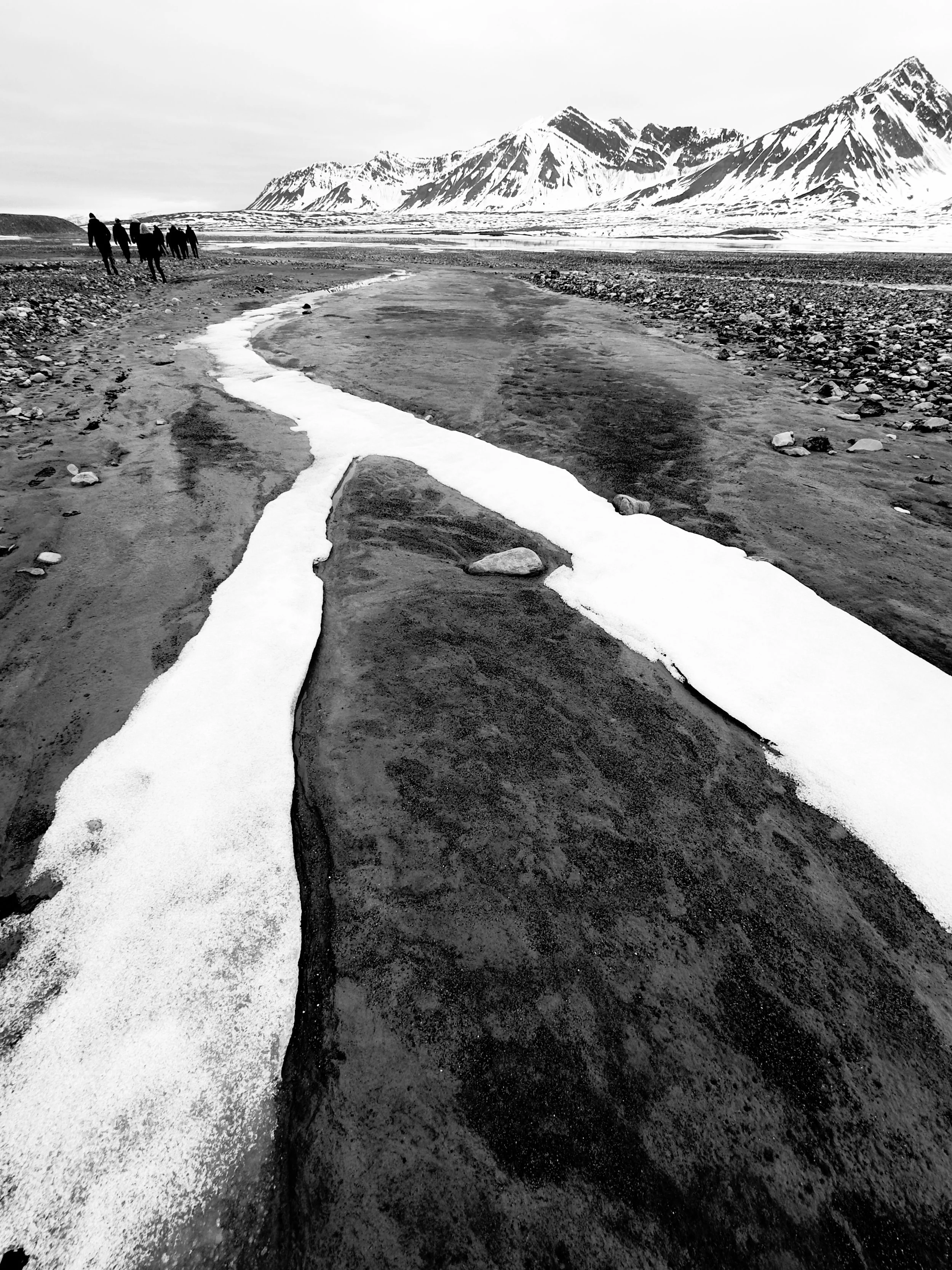 Group of people walking along a riverbed with snow-capped mountains in the background, black and white photograph.