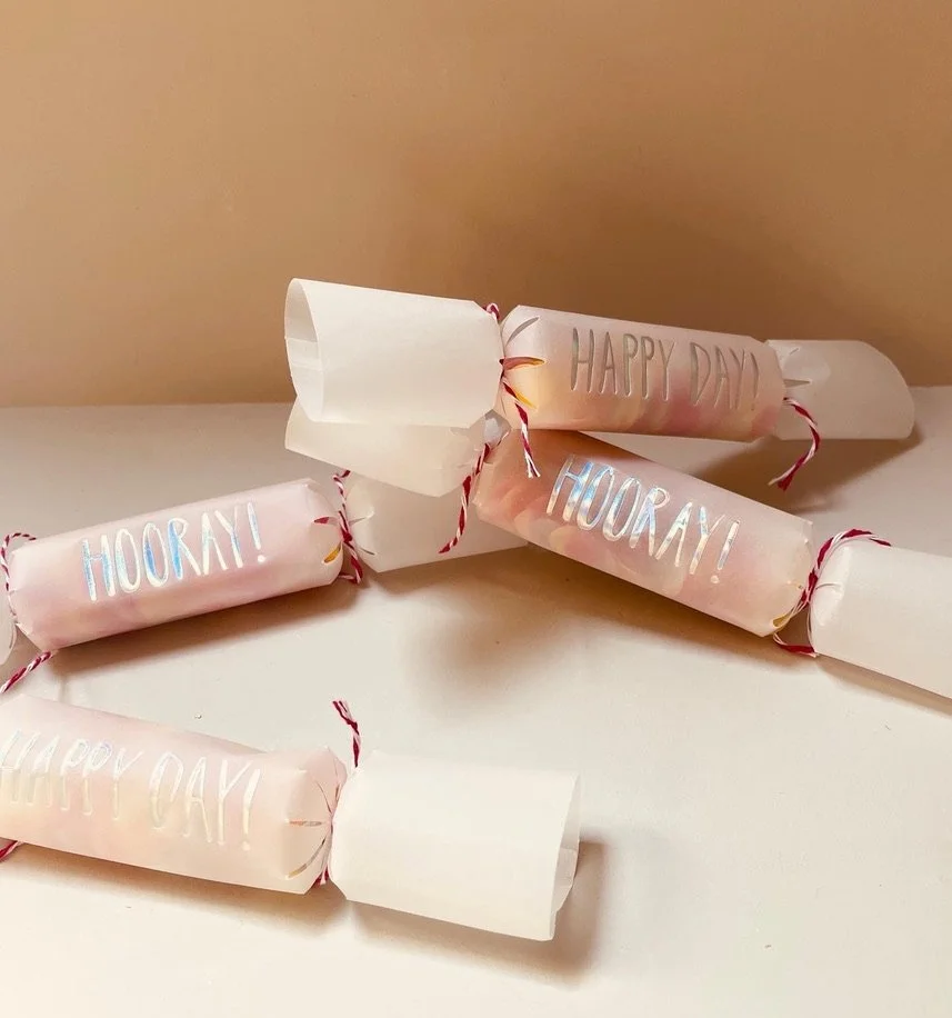 Multiple small firecracker-like objects wrapped in pink paper with 'Hooray!' and 'Happy Day' written on them, decorated with red and white strings, placed on a white surface against a beige background.