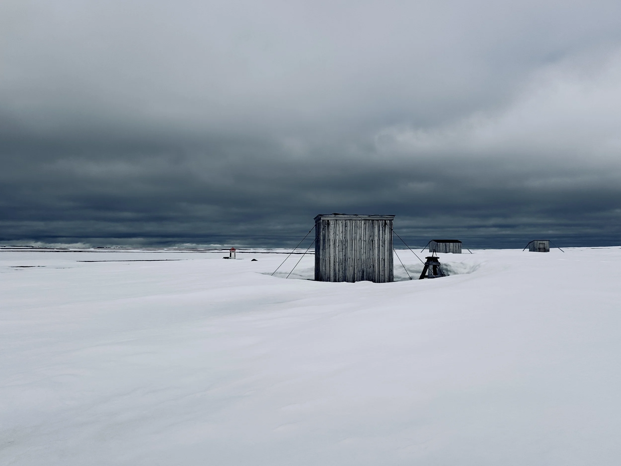 Snow-covered landscape with three wooden cabins under a dark, cloudy sky.