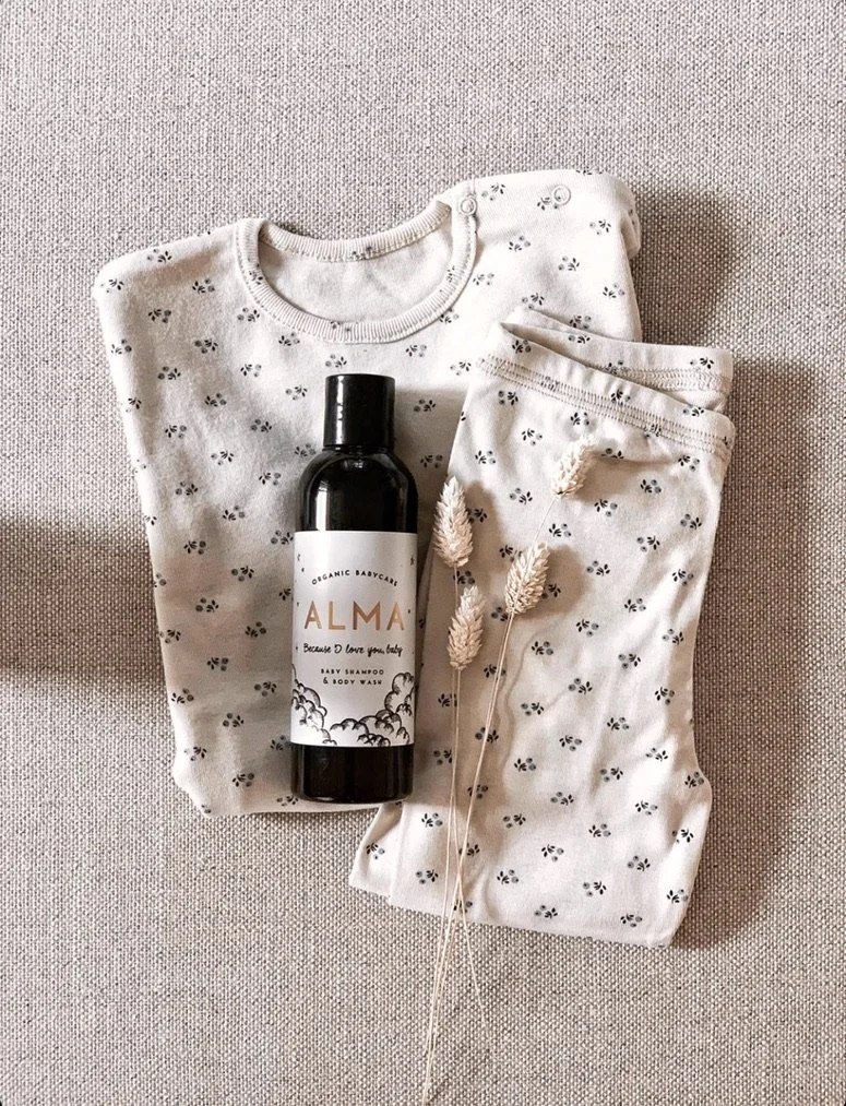 A folded white baby onesie with small black and white flower pattern, a pair of matching baby pants, a black bottle of baby shampoo labeled 'ALMA,' and three decorative beige dried grasses on a neutral-colored fabric surface.