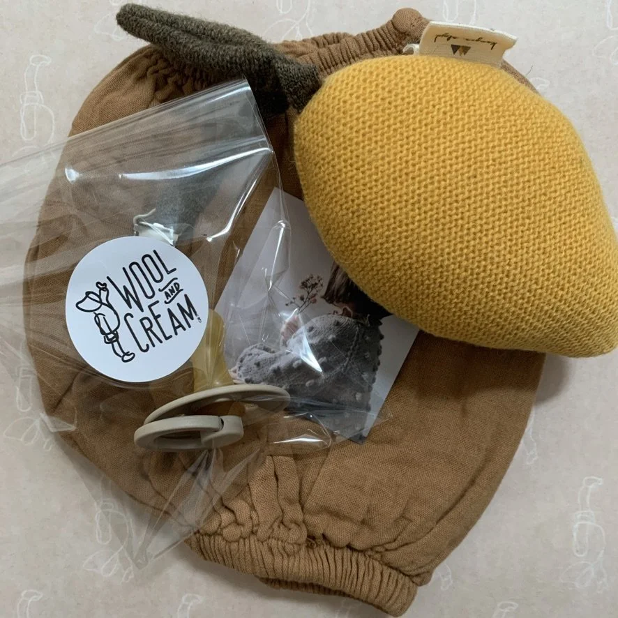 A yellow knitted heart-shaped pouch, a plastic bag with a wool and cream sticker, a pair of beige glasses, and a pair of brown fleece-lined pants