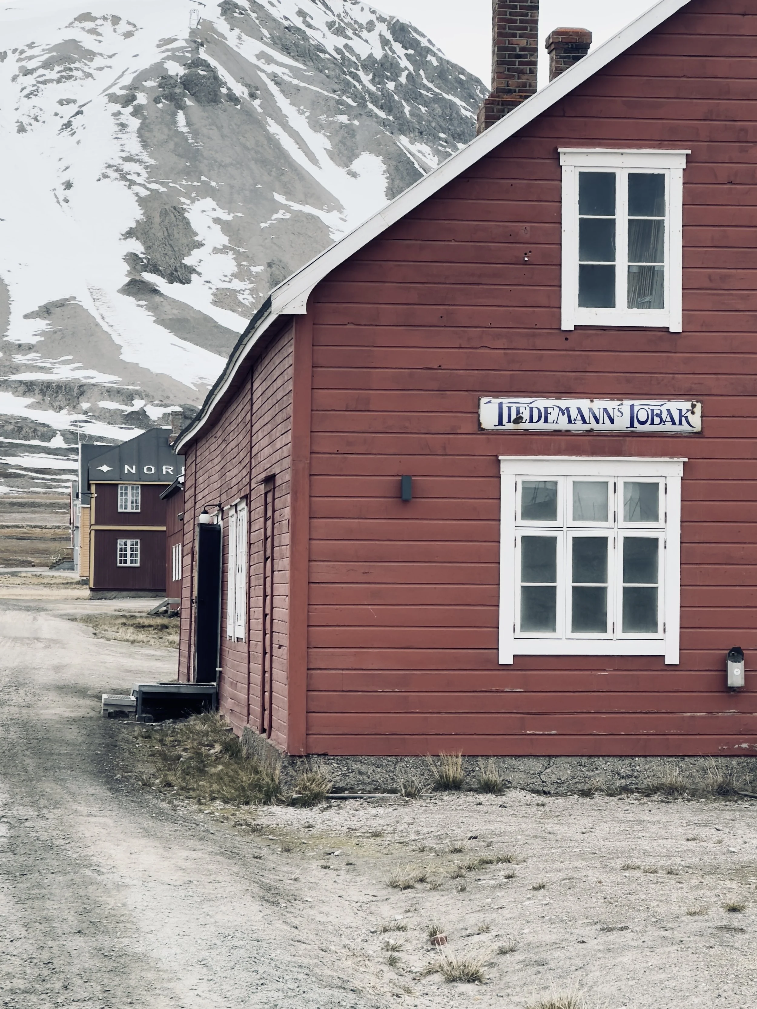 Red wooden building with white-trimmed windows and a sign that reads 'Tiedemann's Tobak', with snow-covered mountains in the background and a dirt road in front.
