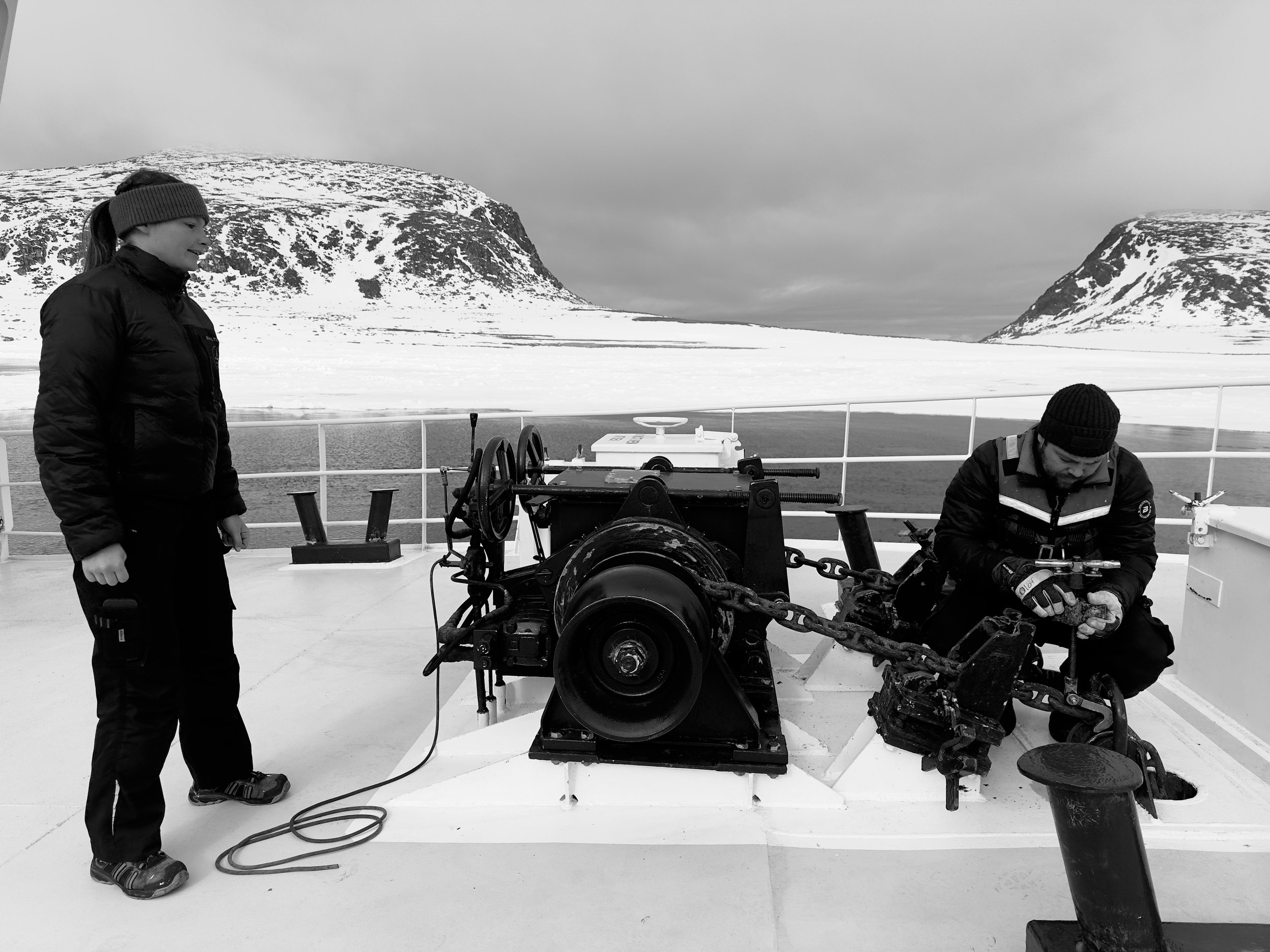 Two people on a boat deck with snow-covered mountains in the background; one person standing, one sitting working on equipment.