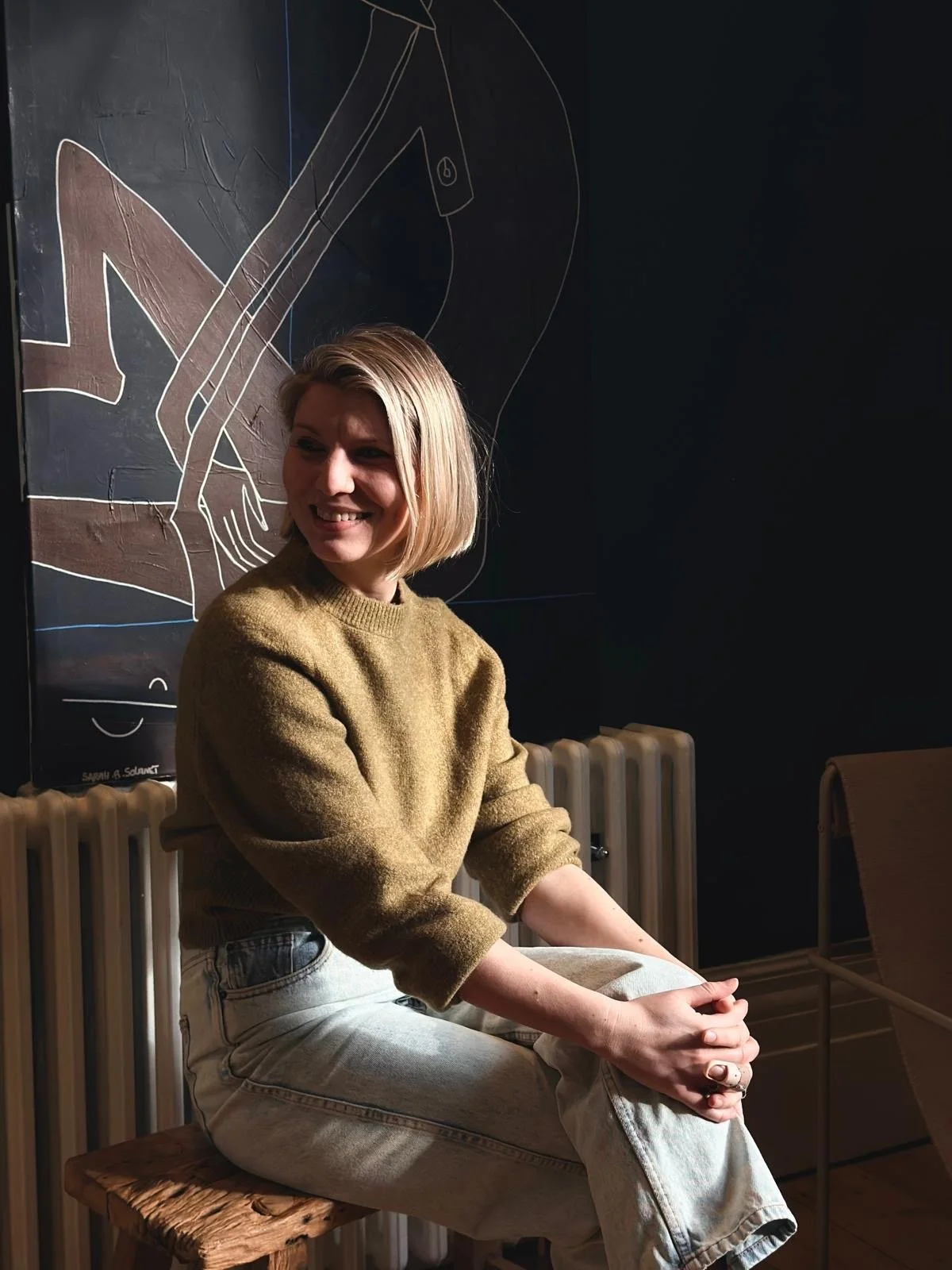 A woman with short blonde hair smiling while sitting on a wooden stool at a cafe or restaurant. She is wearing a brown sweater and light-colored jeans. Behind her is a dark wall with a chalkboard drawing of a person, and a radiator is visible below the drawing.