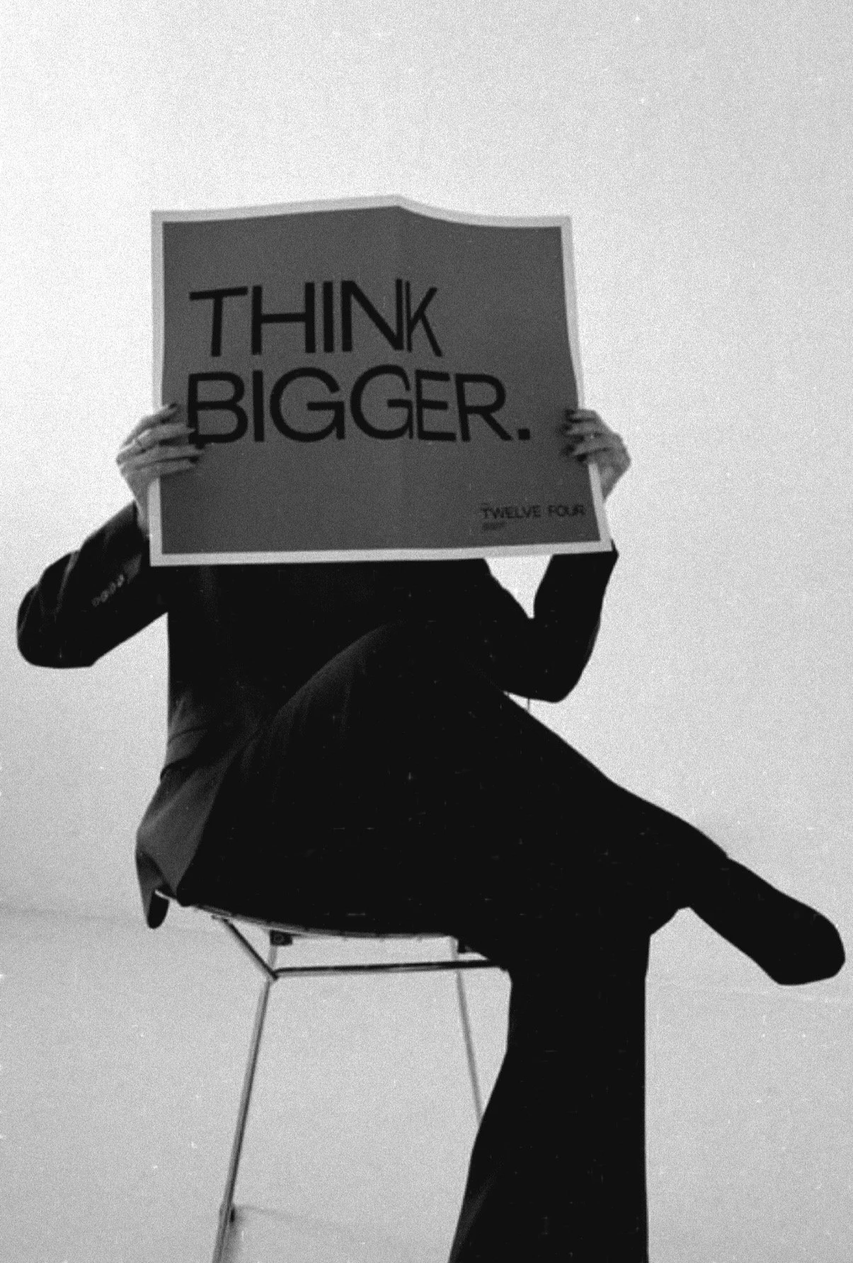 Person in a suit sitting on a chair, holding a sign that says "THINK BIGGER."