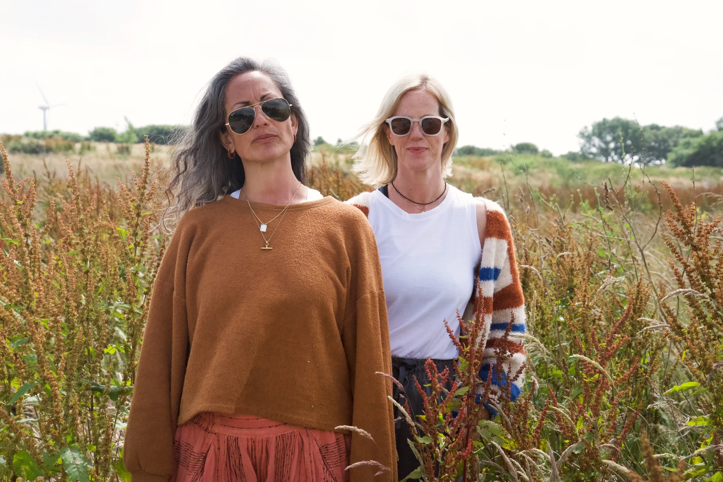 Two women standing in a field of tall plants with a wind turbine in the background. One woman has dark hair, wears sunglasses, a brown sweater, and a pink skirt. The other woman has blonde hair, wears sunglasses, a white tank top, and a cardigan with