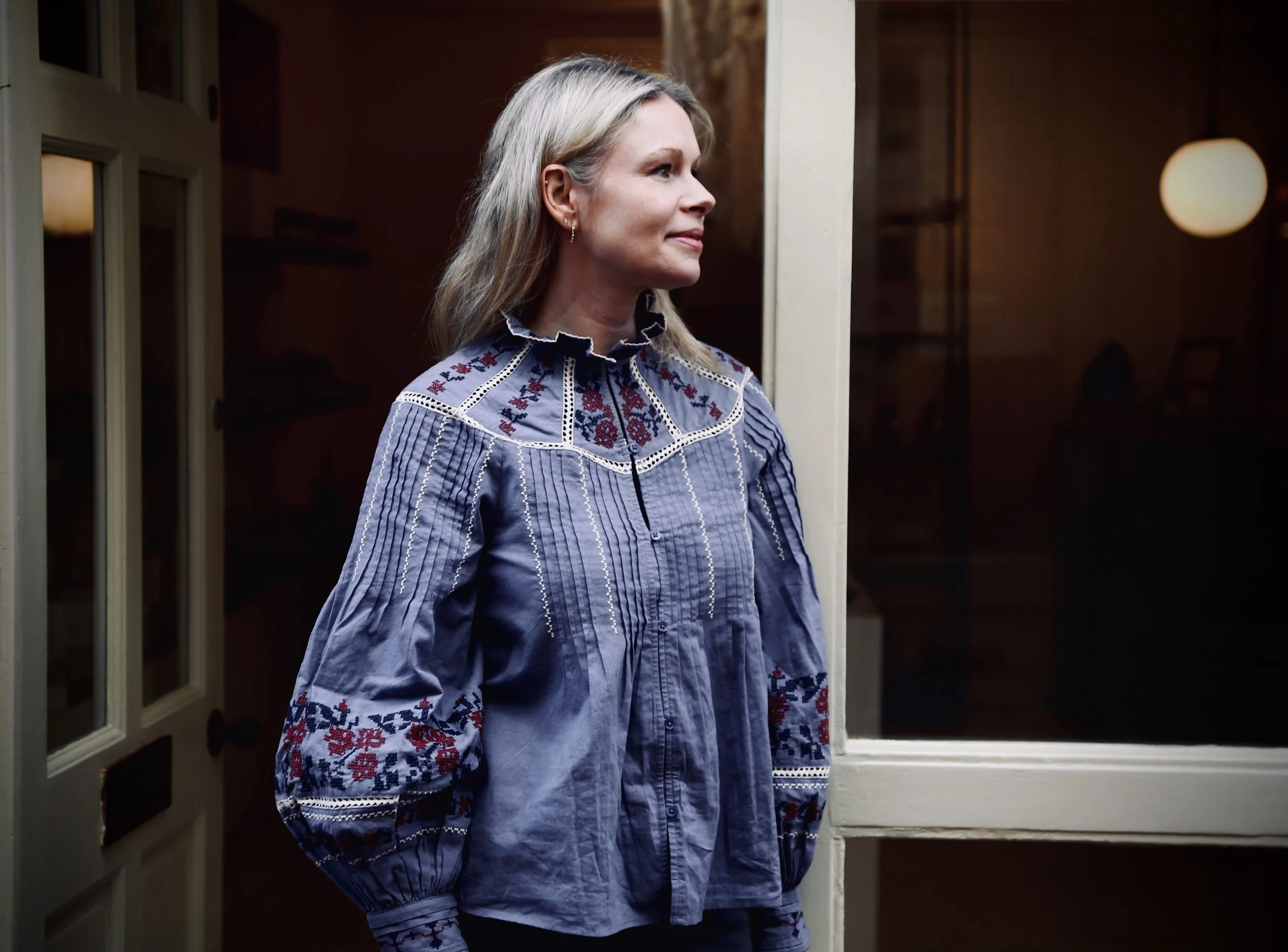 A woman with blonde hair standing in front of a window at night, wearing a blue blouse with embroidered patterns on the shoulders and sleeves.