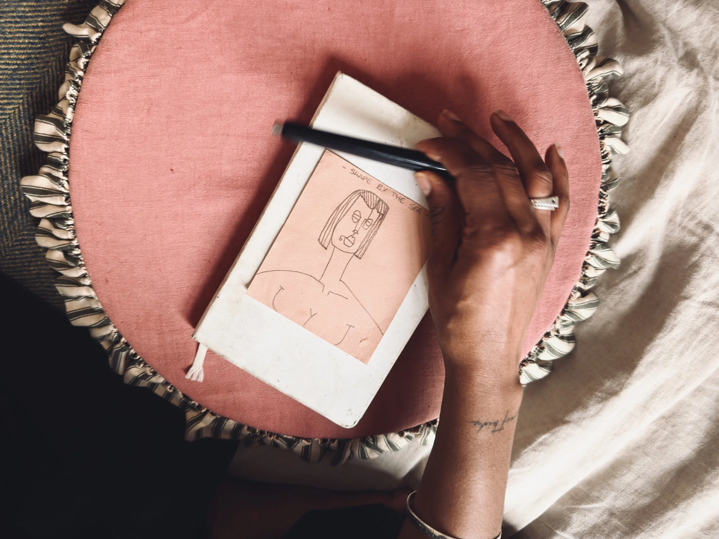 Person sketching a simple line drawing of a person's face and upper body on a tan piece of paper, with the words 'Share by the Sea' at the top, while sitting at a pink, round, fringed table.