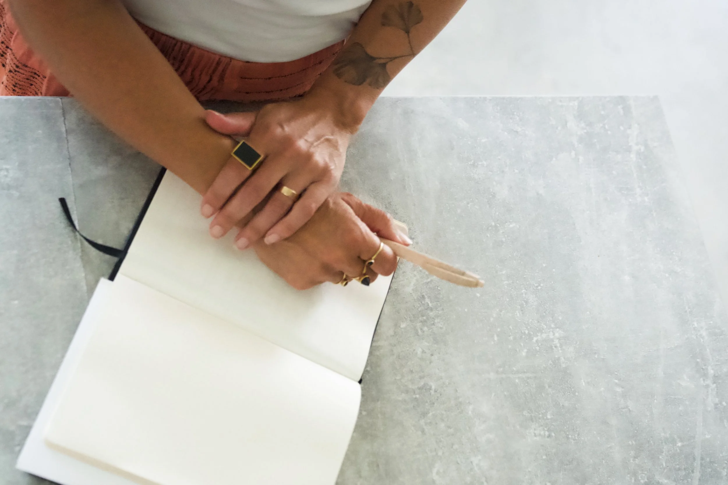Person writing in a blank notebook with a pen on a light gray table, wearing rings and a black ring with a yellow outline, and a tattooed arm.