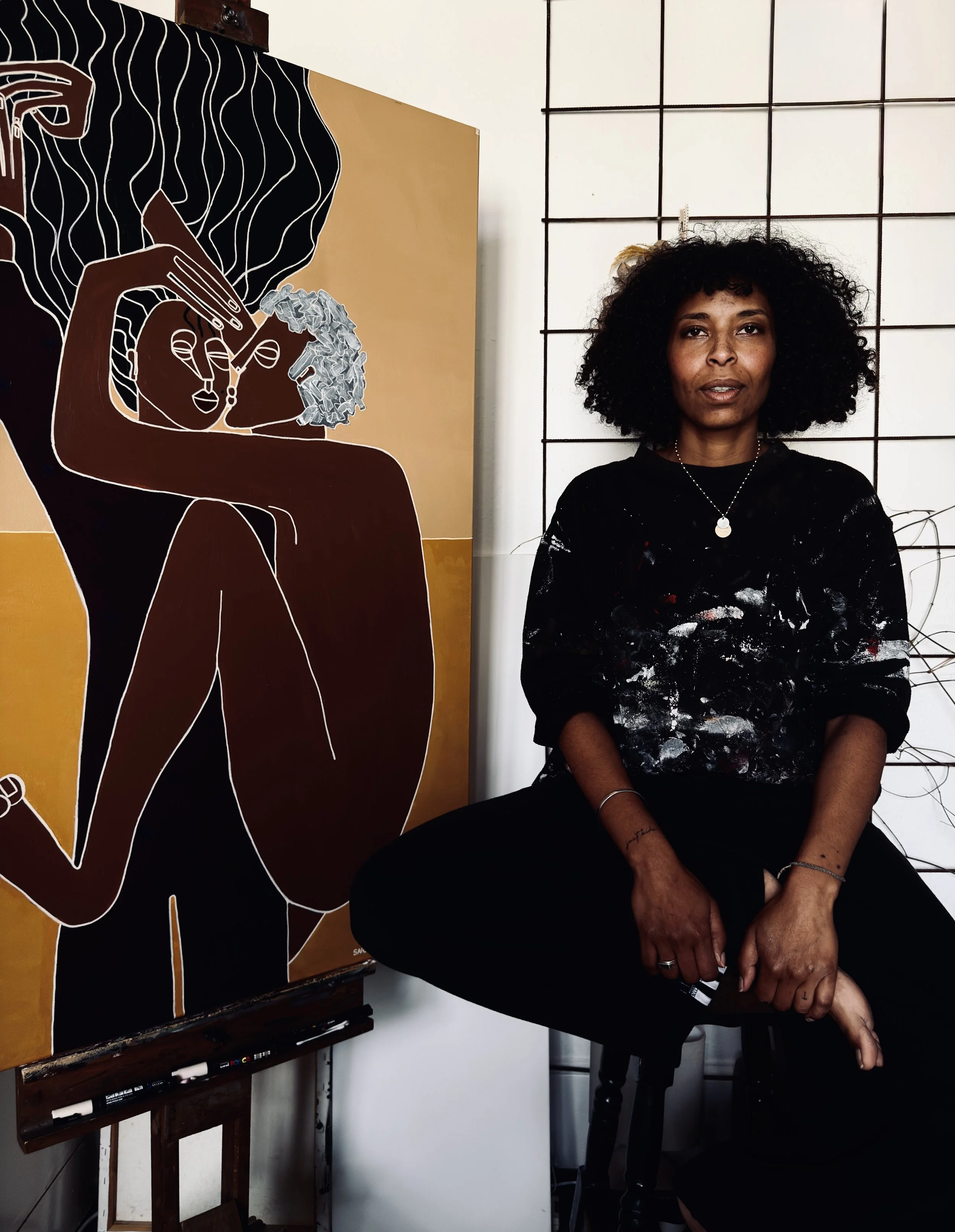 An artist woman sitting on a stool next to her artwork, a stylized painting of a woman with curly hair, who is depicted in abstract black, white, and yellow with her eyes closed, holding a flower near her face.