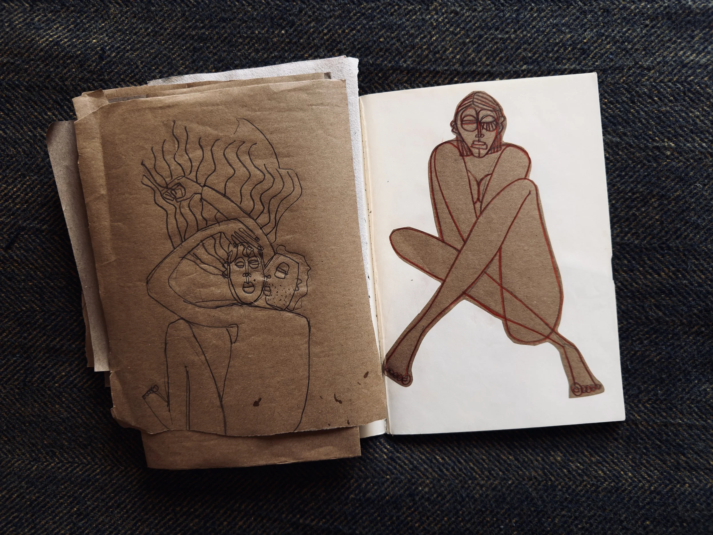 Open sketchbook with two drawings of a woman. The left drawing is a line sketch on brown paper, showing a woman with wavy hair, holding her head, and a face or mask beside her. The right drawing is a colored outline on white paper, depicting a woman sitting with crossed legs, her arms wrapped around her knees, with short hair and glasses.