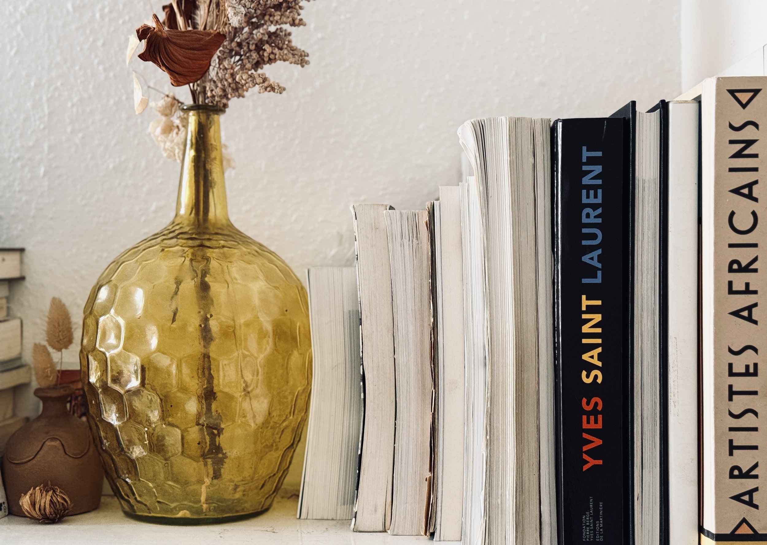 A yellow textured glass vase with dried flowers and plants, arranged on a shelf next to a variety of stacked books.