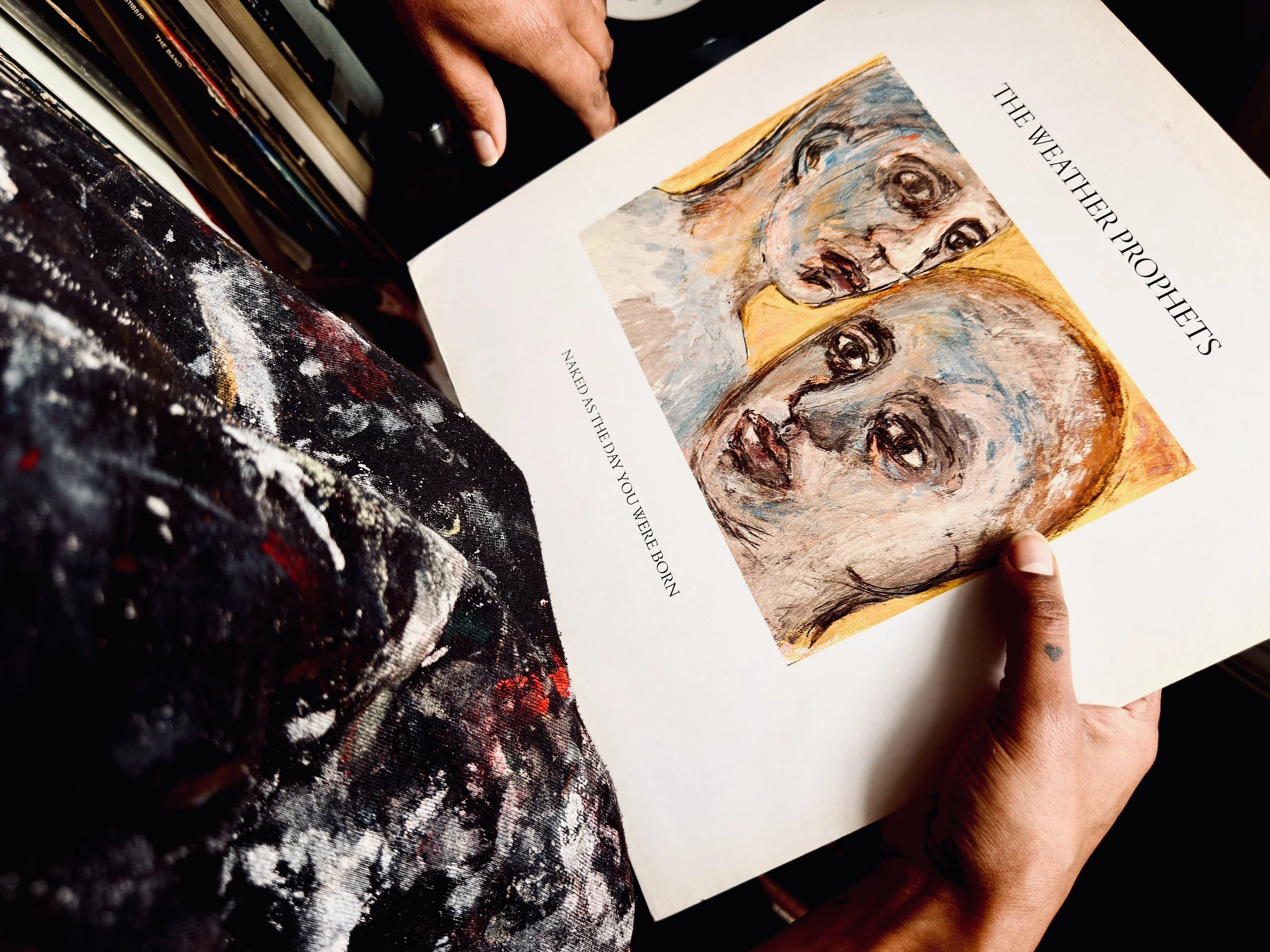 Person holding an open book showing an illustration of two faces with the title "The Weather Prophets" and a quote, with a painterly cluttered apron visible in the foreground.