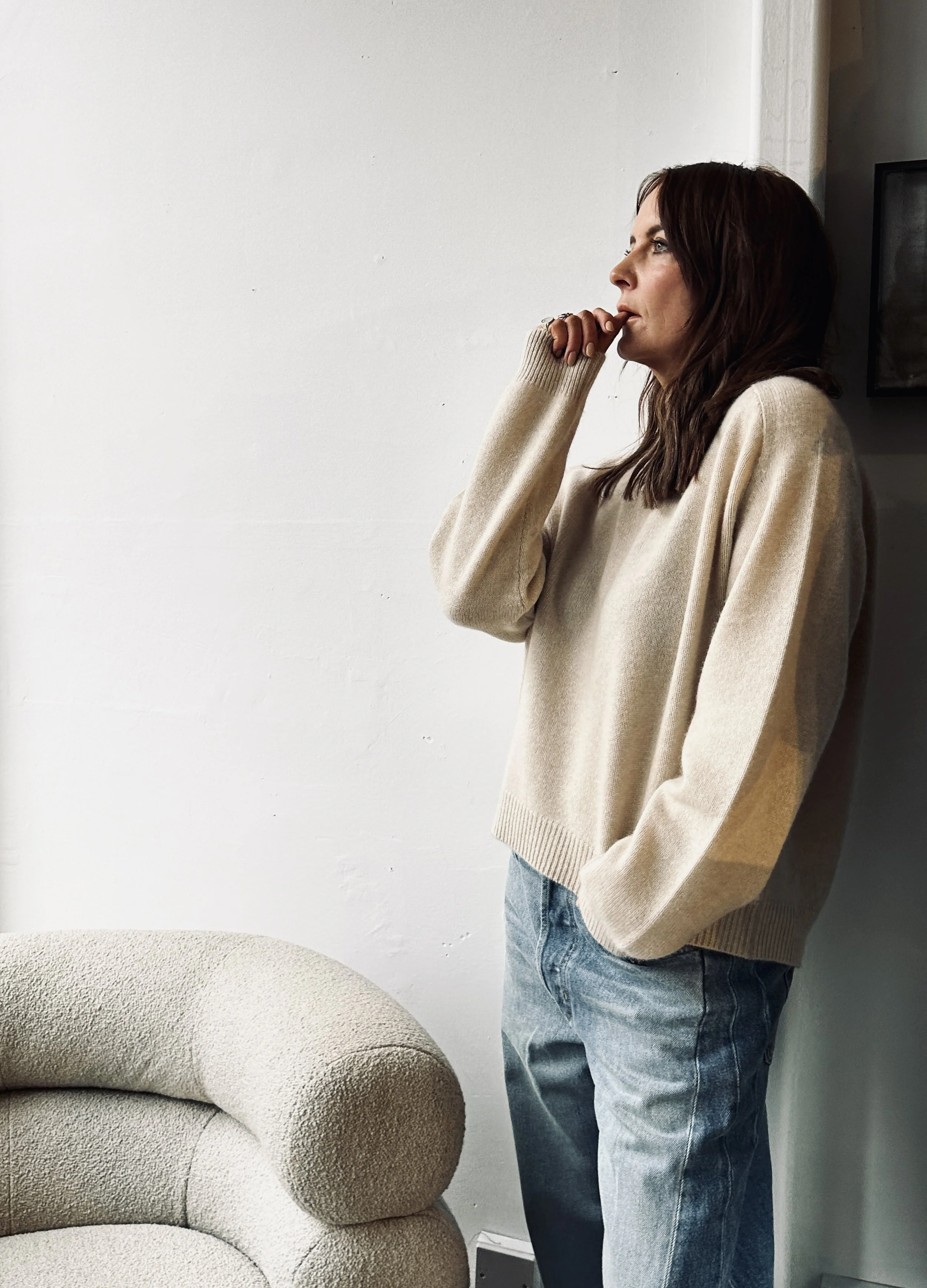 A woman with dark brown hair wearing a beige sweater and blue jeans standing with her hand on her chin, looking off to the side, in a room with a white wall and a beige armchair.