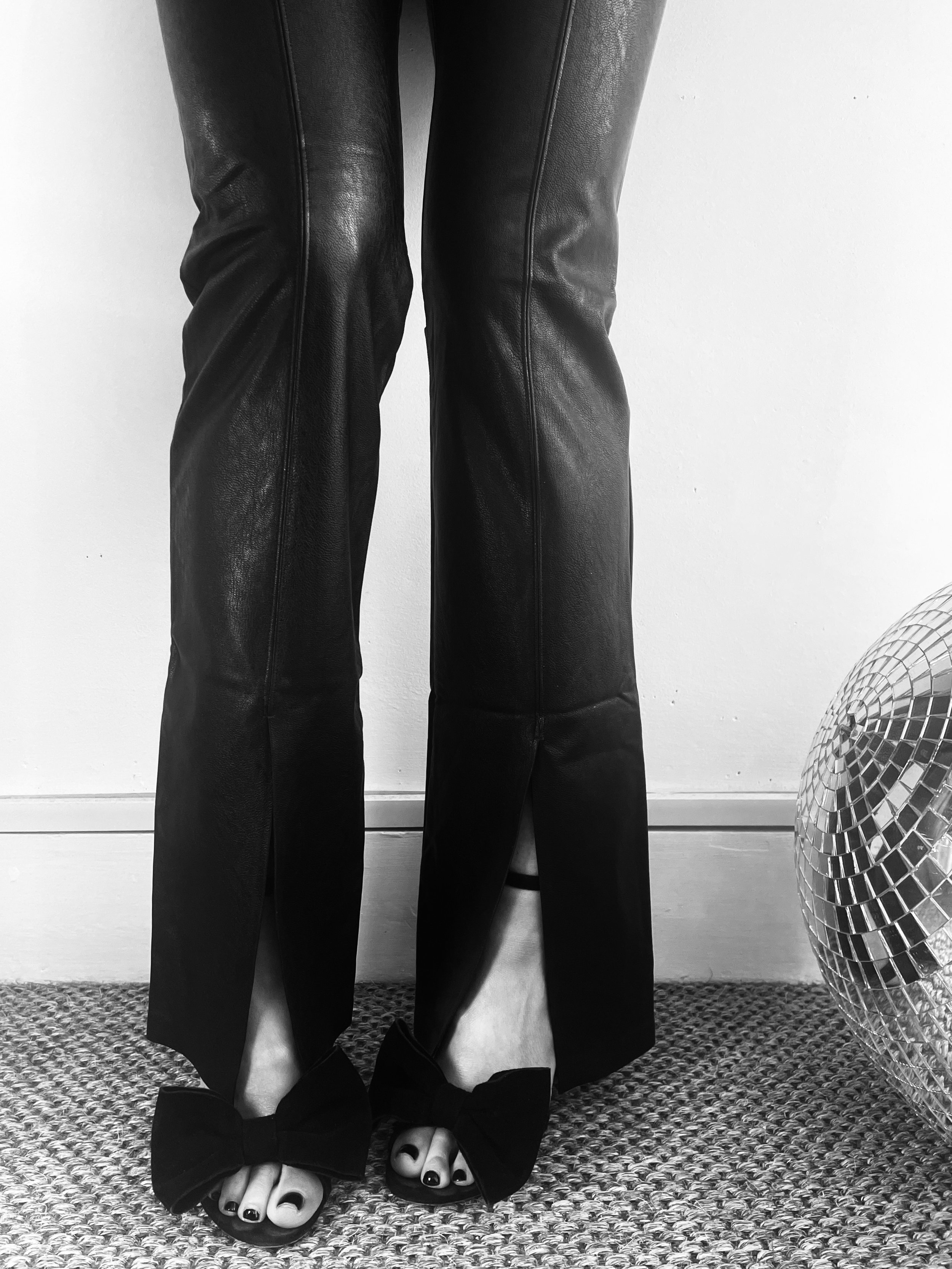 Black leather pants with black high-heeled shoes featuring large bow decor, showing painted toenails, next to a disco ball on a textured carpet.