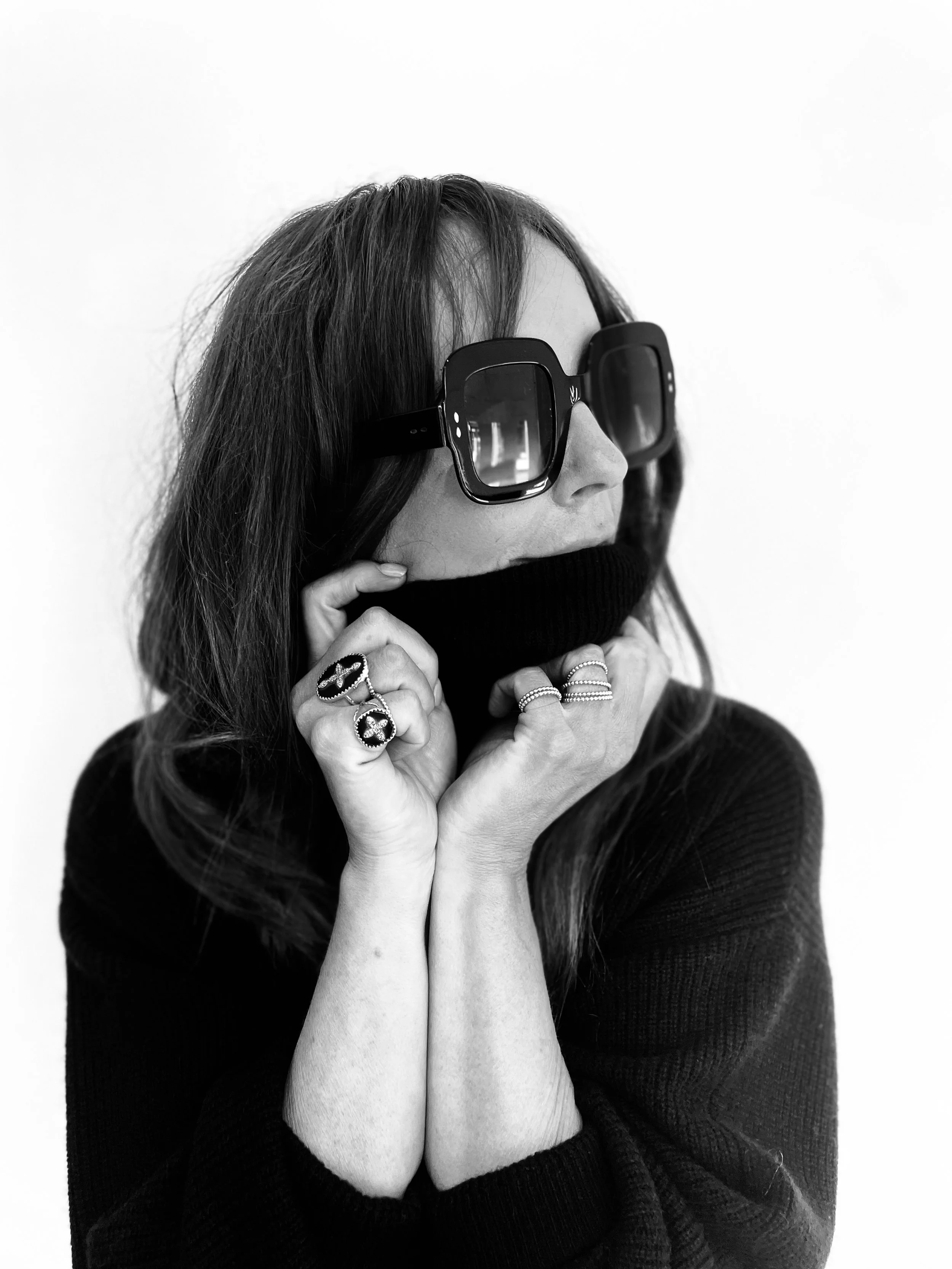 A woman wearing large sunglasses, a dark sweater, and multiple rings, holding a scarf or turtleneck partially covering her face, with a plain white background.