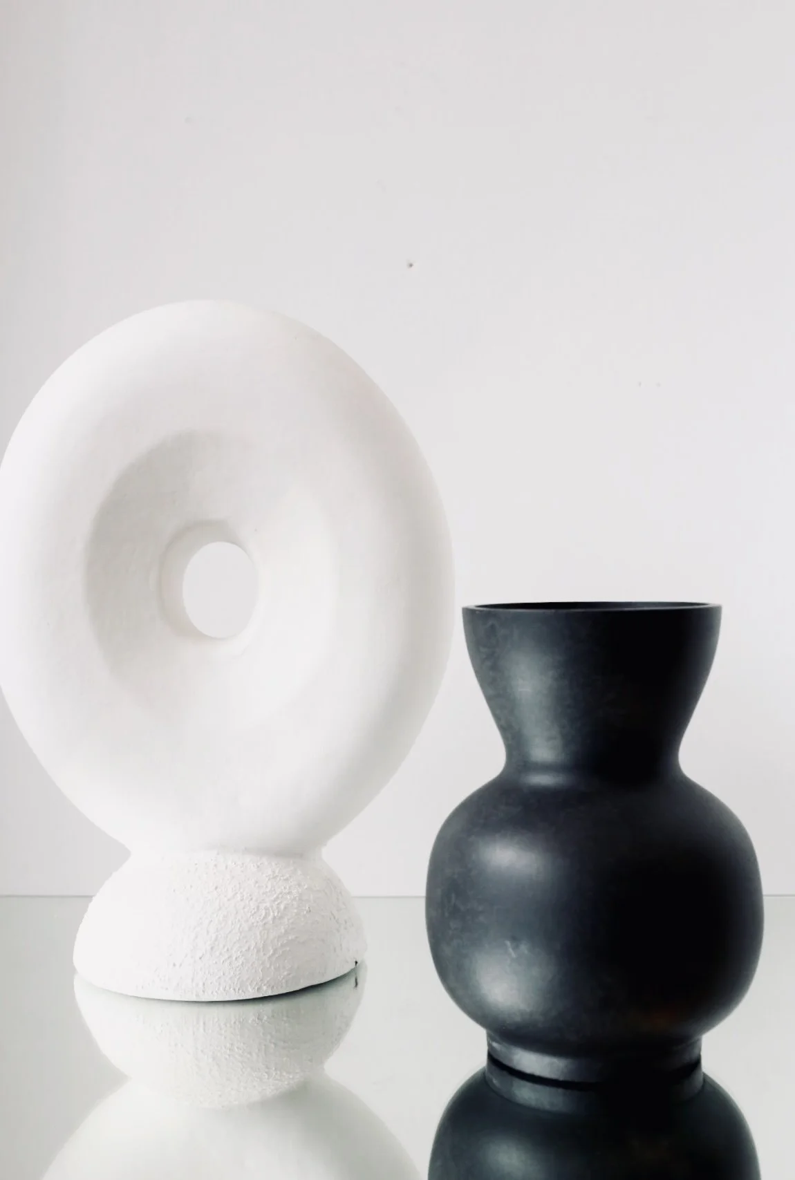 Two minimalist vases on a reflective surface against a plain white background, one black and bulbous, the other white and disc-shaped.