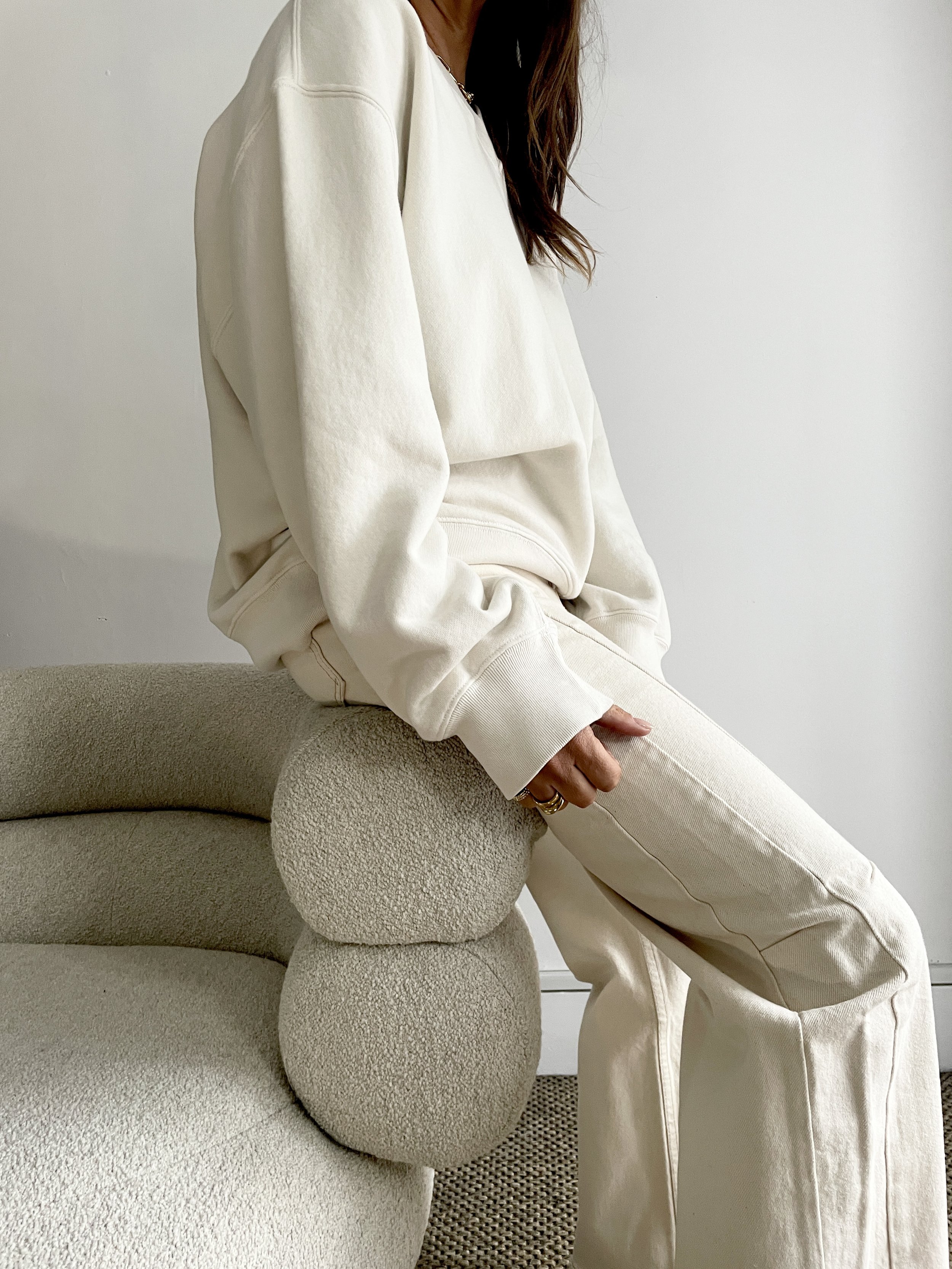 Person sitting on a beige textured sofa with two rounded cushions under their arm, wearing a cream-colored oversized sweatshirt and pants, with long dark hair and rings on their hand, against a plain white wall.