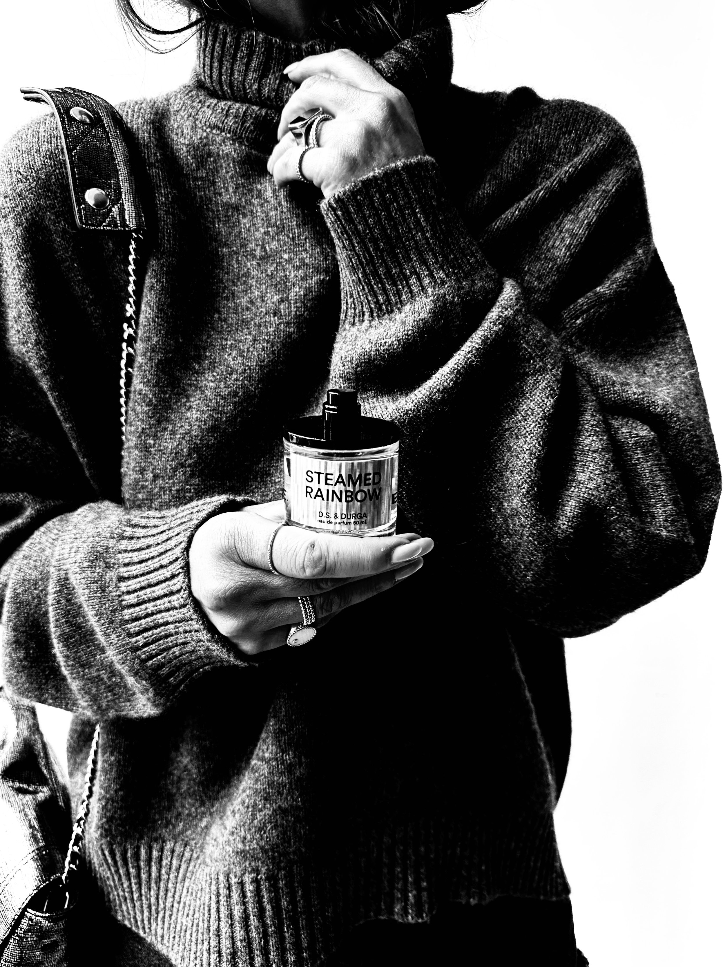 Person in a knitted sweater holding a perfume bottle labeled 'Steamed Rainbow' close to their chest, with a hand touching their face.