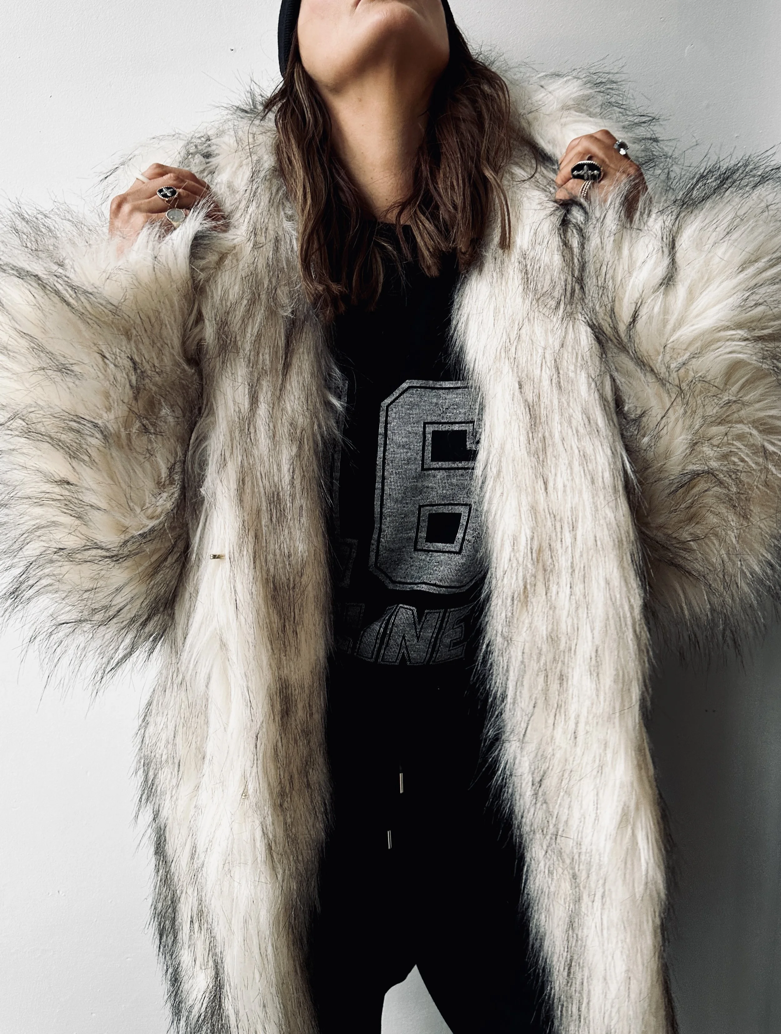 Person wearing a large, fluffy, cream-colored fur coat with black accents, holding the coat collar with hands, standing against a plain white wall.