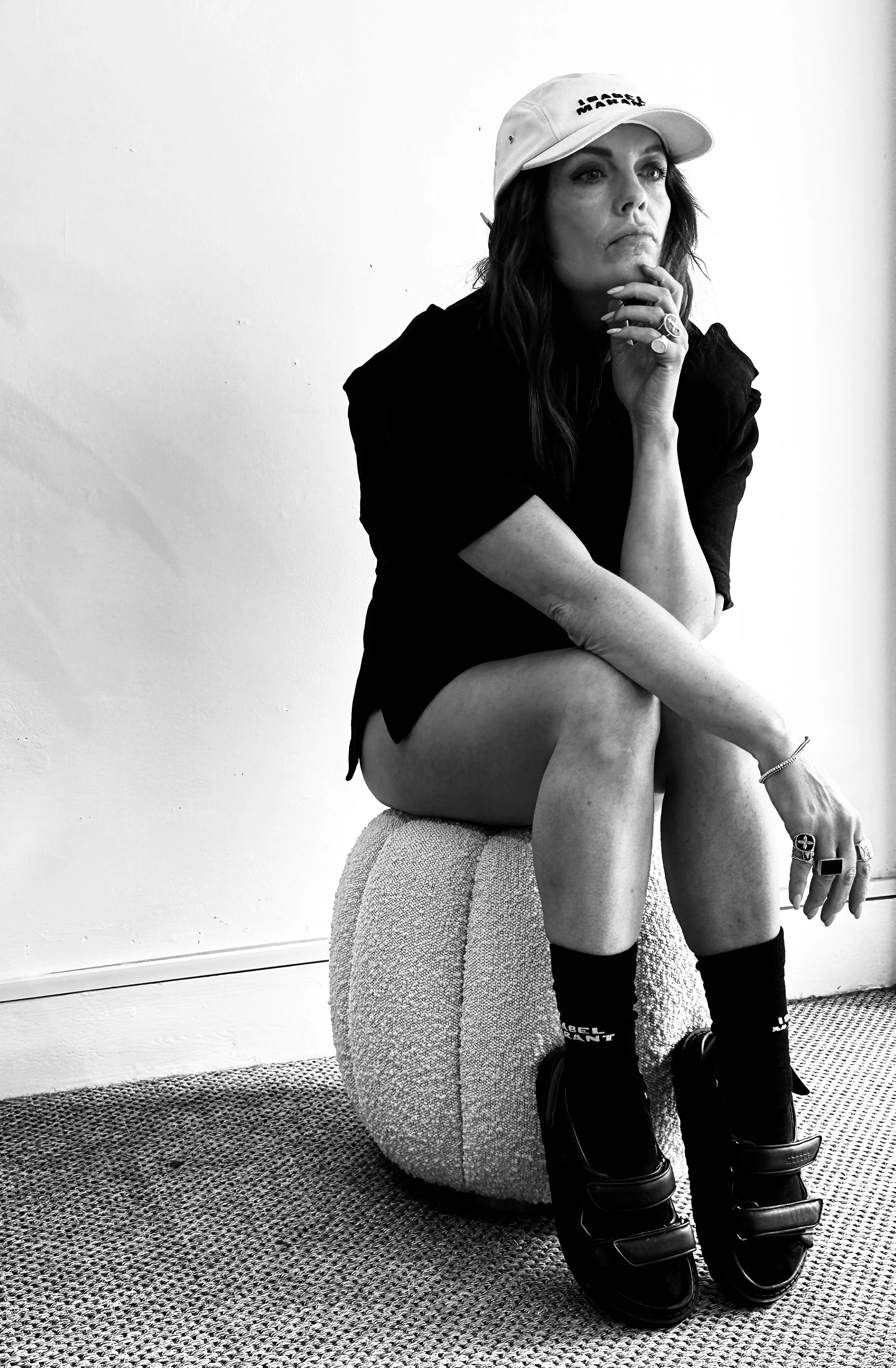 A woman sitting on a textured round ottoman, wearing a baseball cap, dark loose shirt, socks, and sandals, with her hand on her chin and looking to the side, in a room with plain walls.