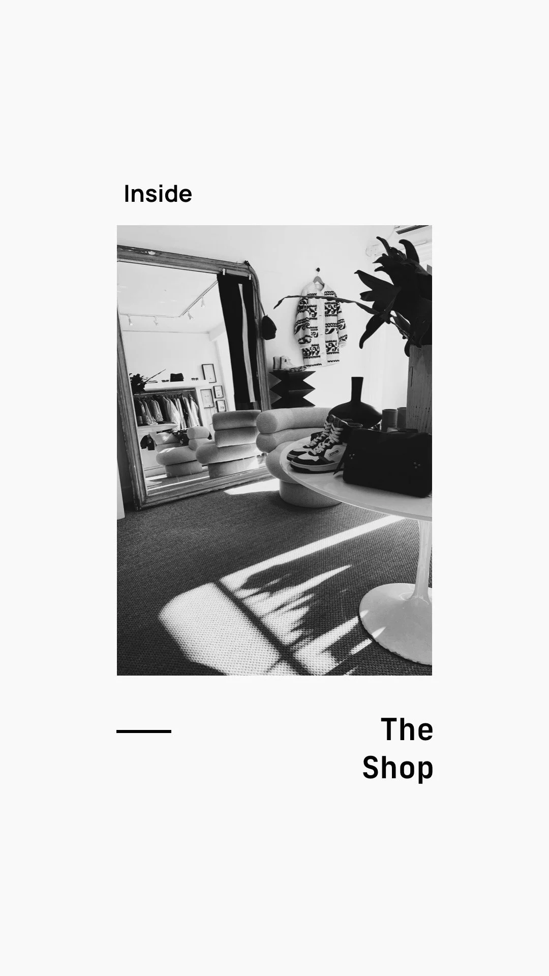 A black and white photo of a stylish interior space with a modern round chair, a table with shoes and a purse, and a large mirror reflecting a clothing rack and decor in a boutique shop.