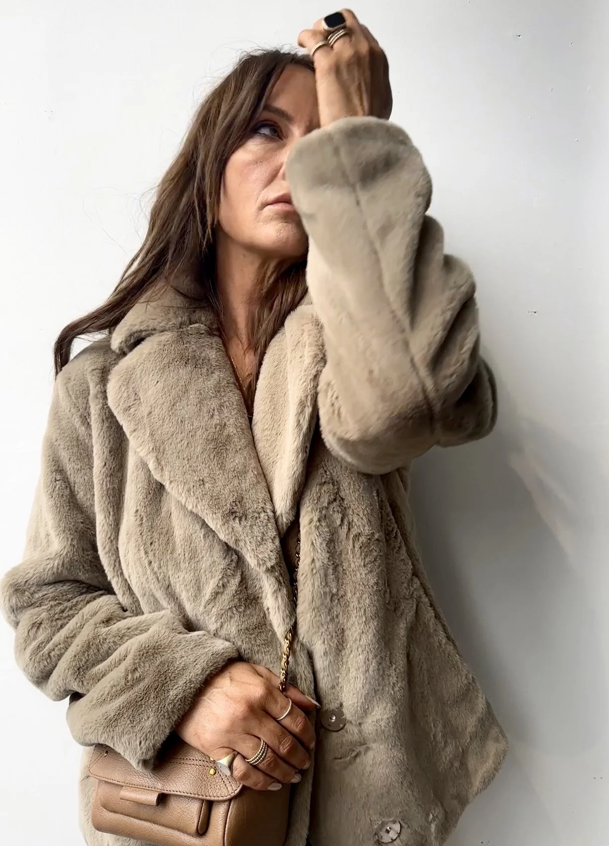 A woman with long brown hair wearing a beige fur coat and carrying a small beige purse, standing against a plain white wall.