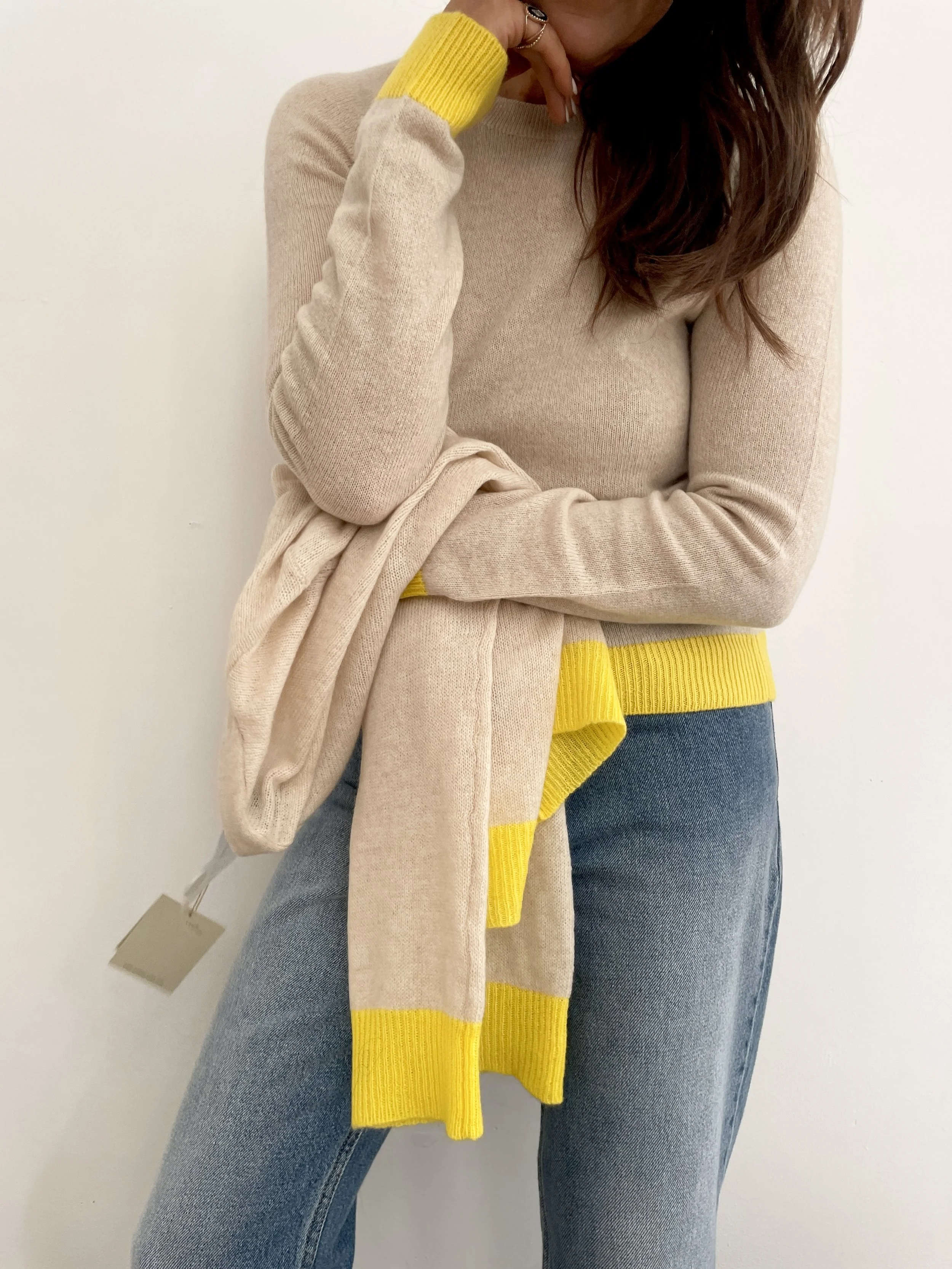 A woman wearing a beige sweater with yellow accents on the cuffs and waist, paired with blue jeans, is holding a matching beige and yellow scarf. She stands against a plain white background, with her face partially cropped out.