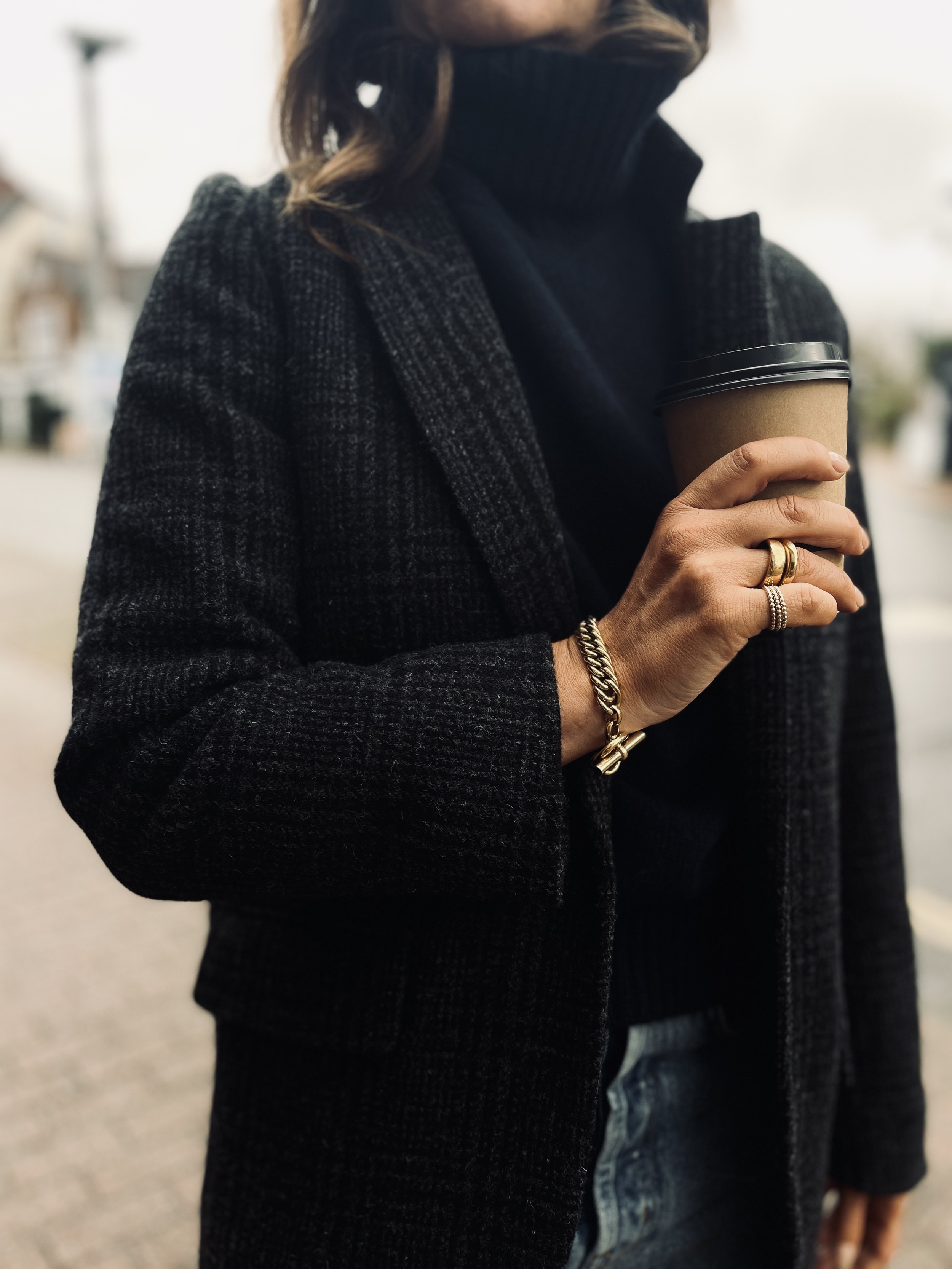 A woman wearing a dark grey plaid coat and black turtleneck sweater, holding a disposable coffee cup, outdoors on a cloudy day.