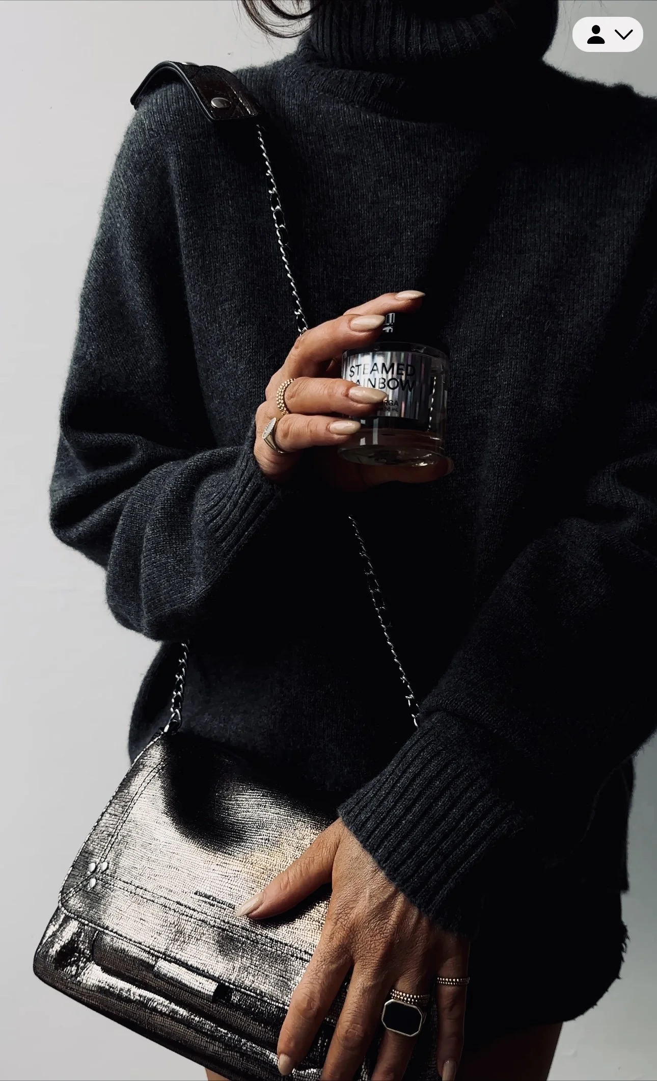 Person dressed in a black sweater holding a black scented candle and wearing rings, with a metallic silver crossbody bag.