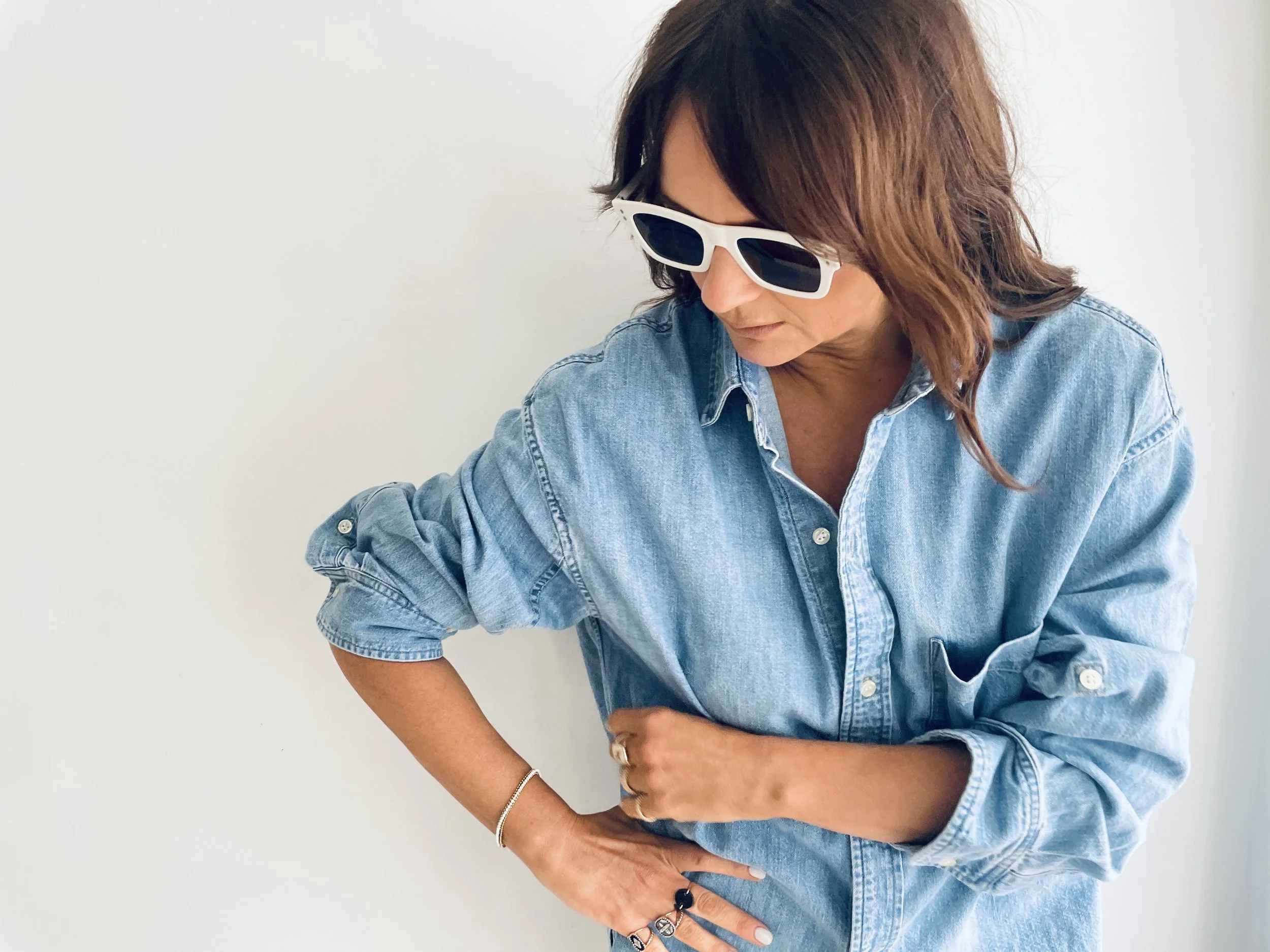 Woman with brown hair wearing white sunglasses and a denim shirt, looking down with her right hand on her waist.