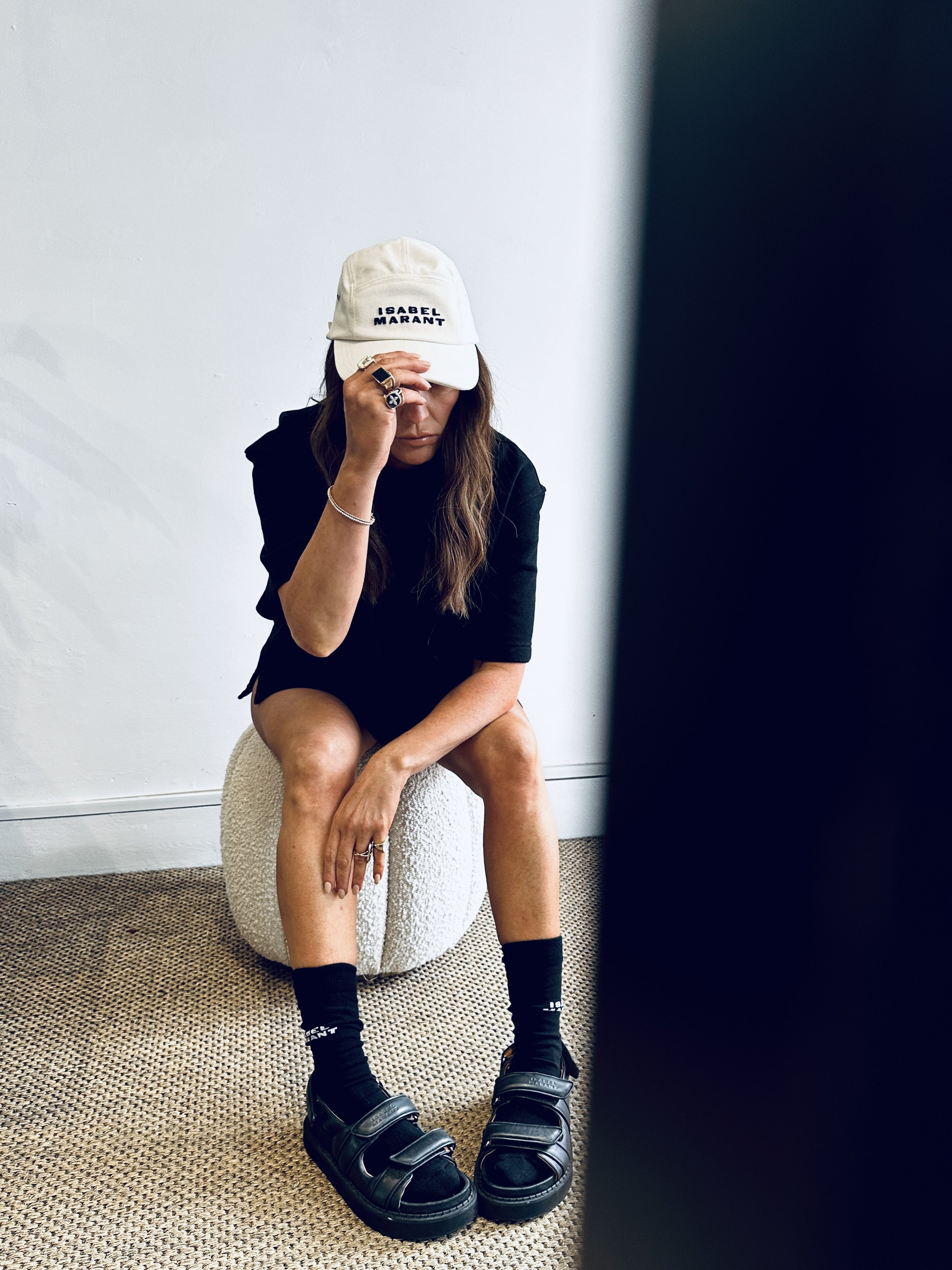 Woman sitting on textured ottoman, wearing a white cap with 'ISABEL MARANT' written on it, black outfit, black socks, and chunky black sandals, holding her forehead with her right hand, in a minimalist room with white wall and beige carpet.