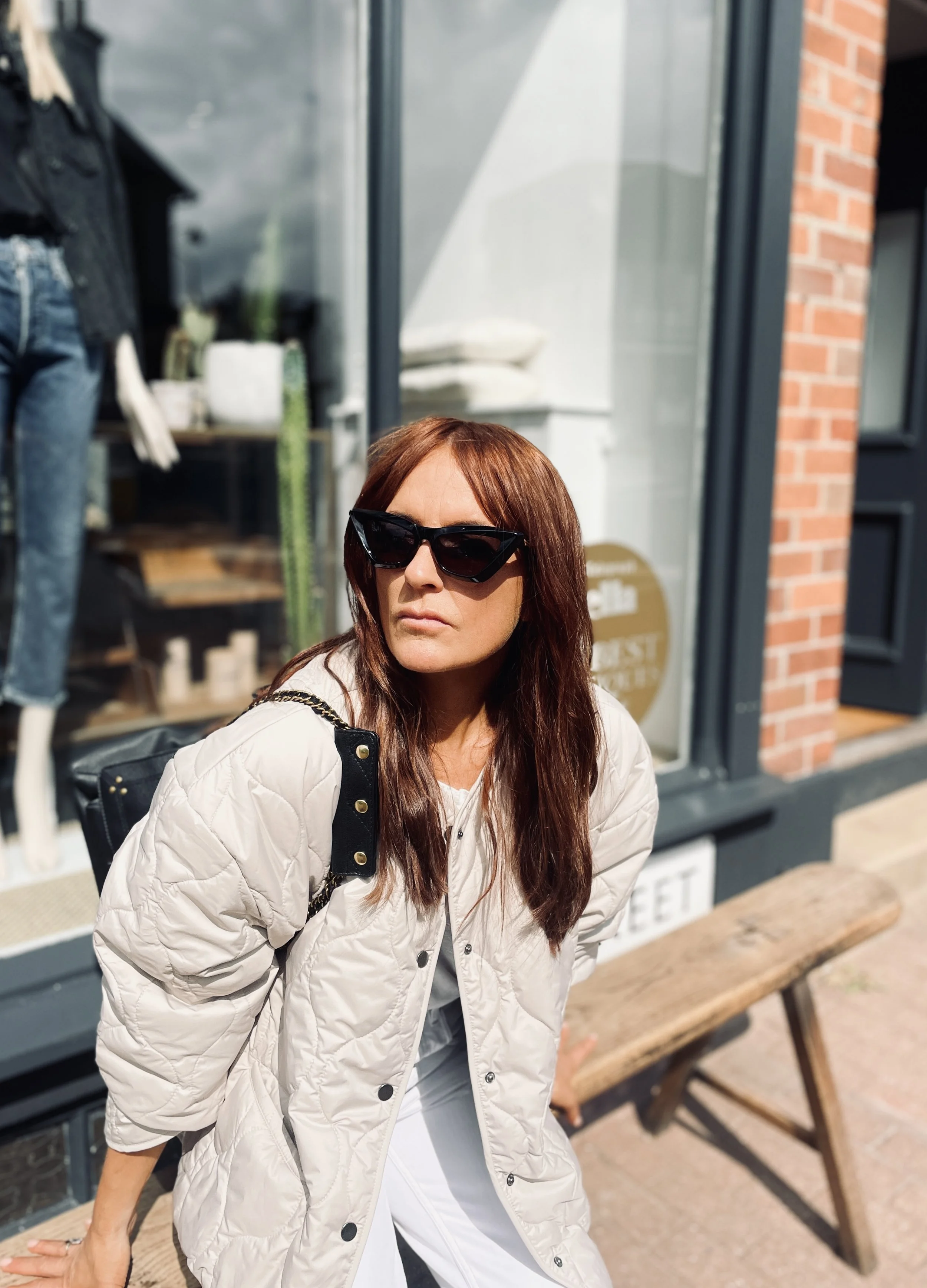 A woman with long brown hair wearing black sunglasses, a cream-colored quilted jacket, and white pants, sitting on a wooden bench outside a storefront with a brick and glass facade.