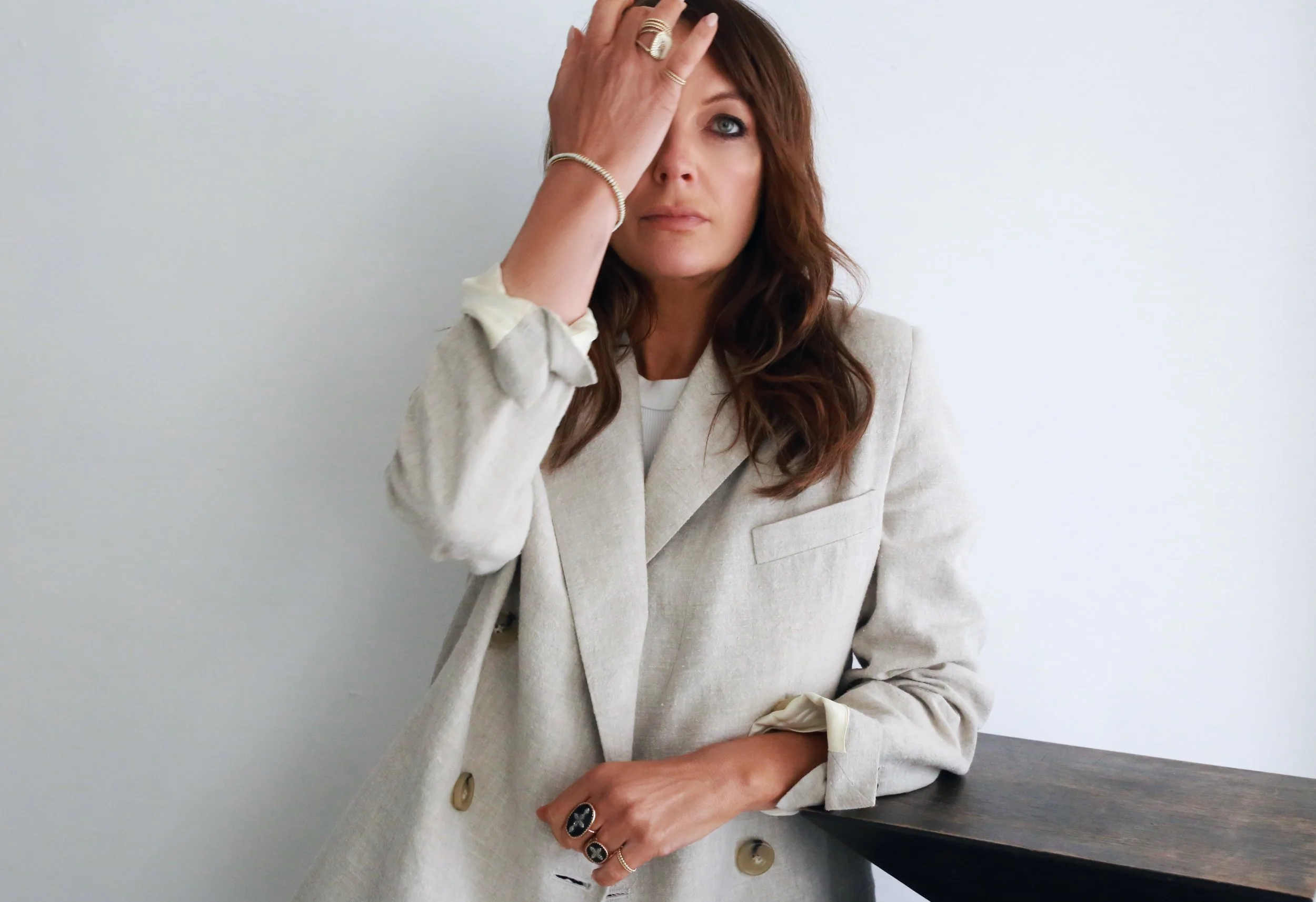 A woman with brown wavy hair wearing a beige blazer, resting her left hand on a dark wooden table, with her right hand covering her right eye, standing against a plain white wall.