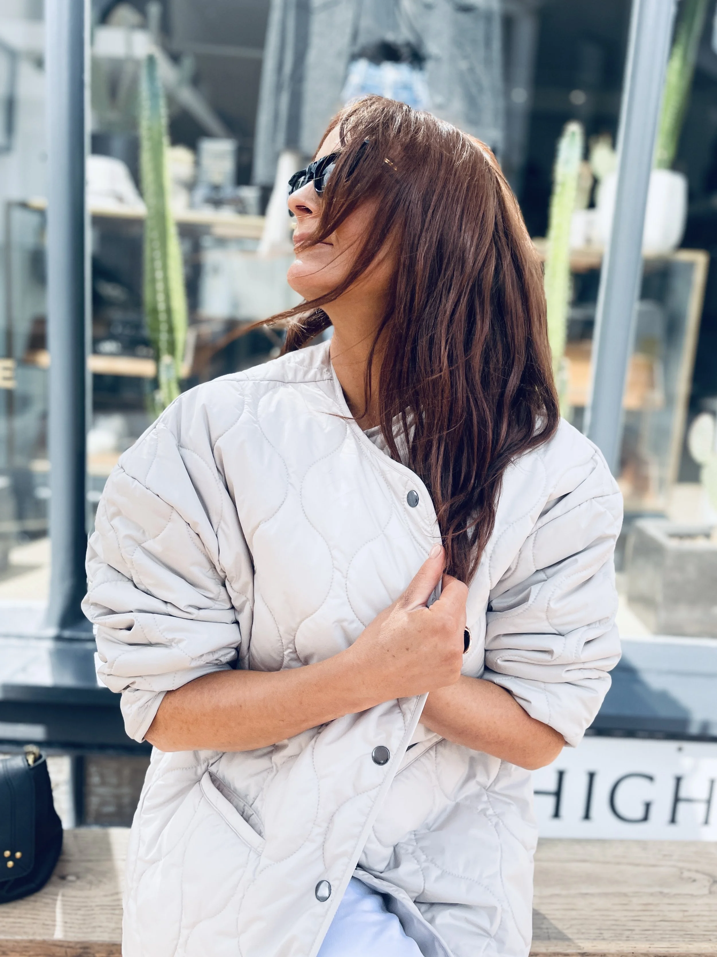 A woman with long brown hair and sunglasses, wearing a beige quilted jacket, standing outdoors with a glass window and cacti in the background.