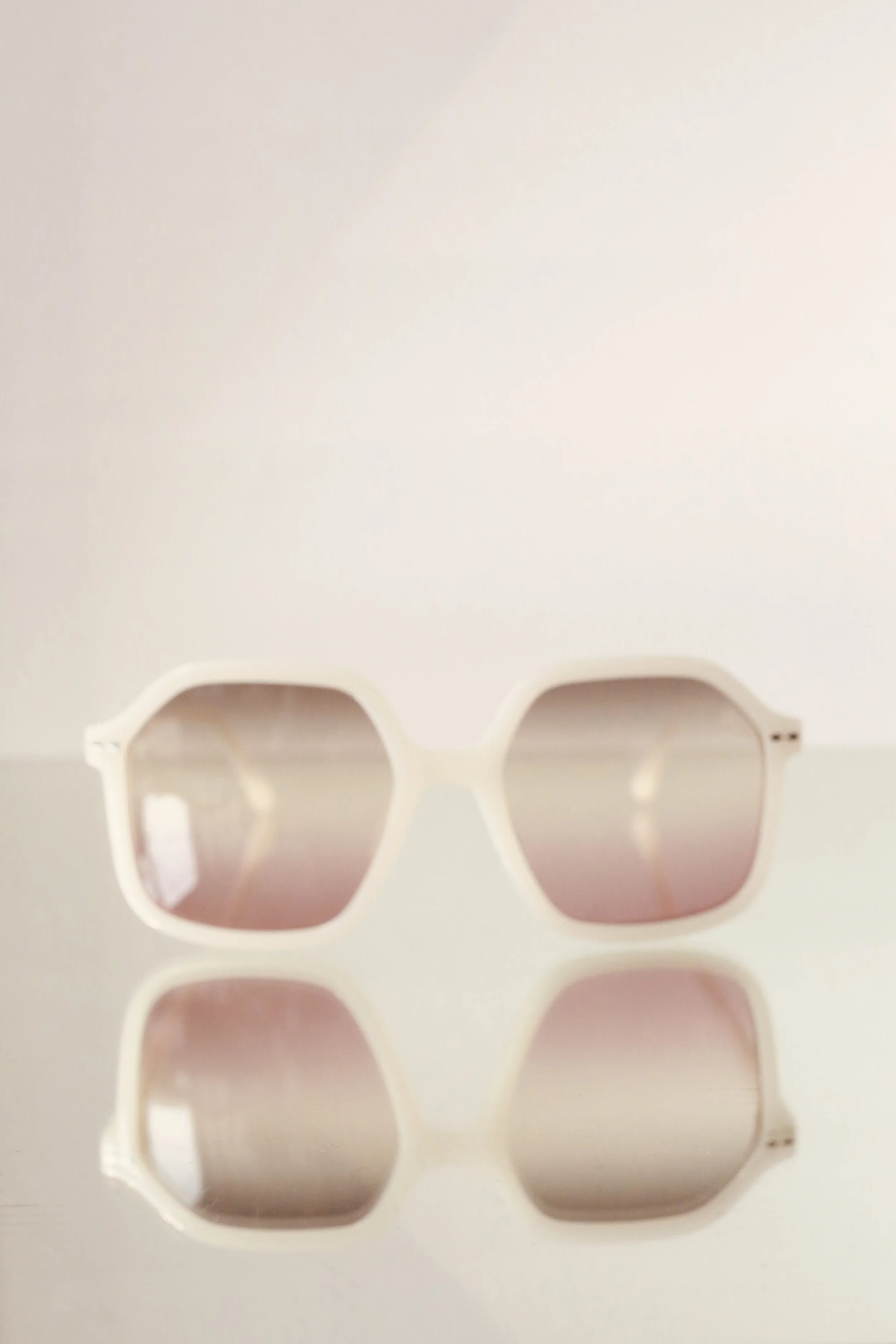 White sunglasses with gradient lenses resting on a reflective surface against a plain white background.