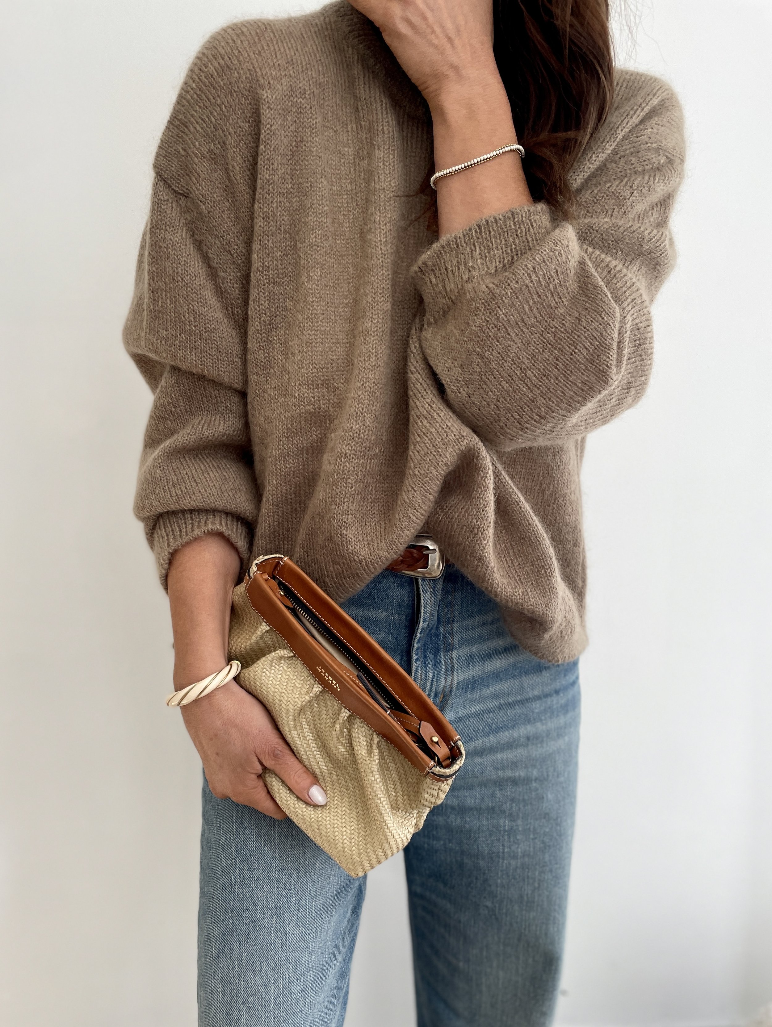 Woman in beige sweater holding a tan clutch bag and wearing jeans, standing against a white wall.