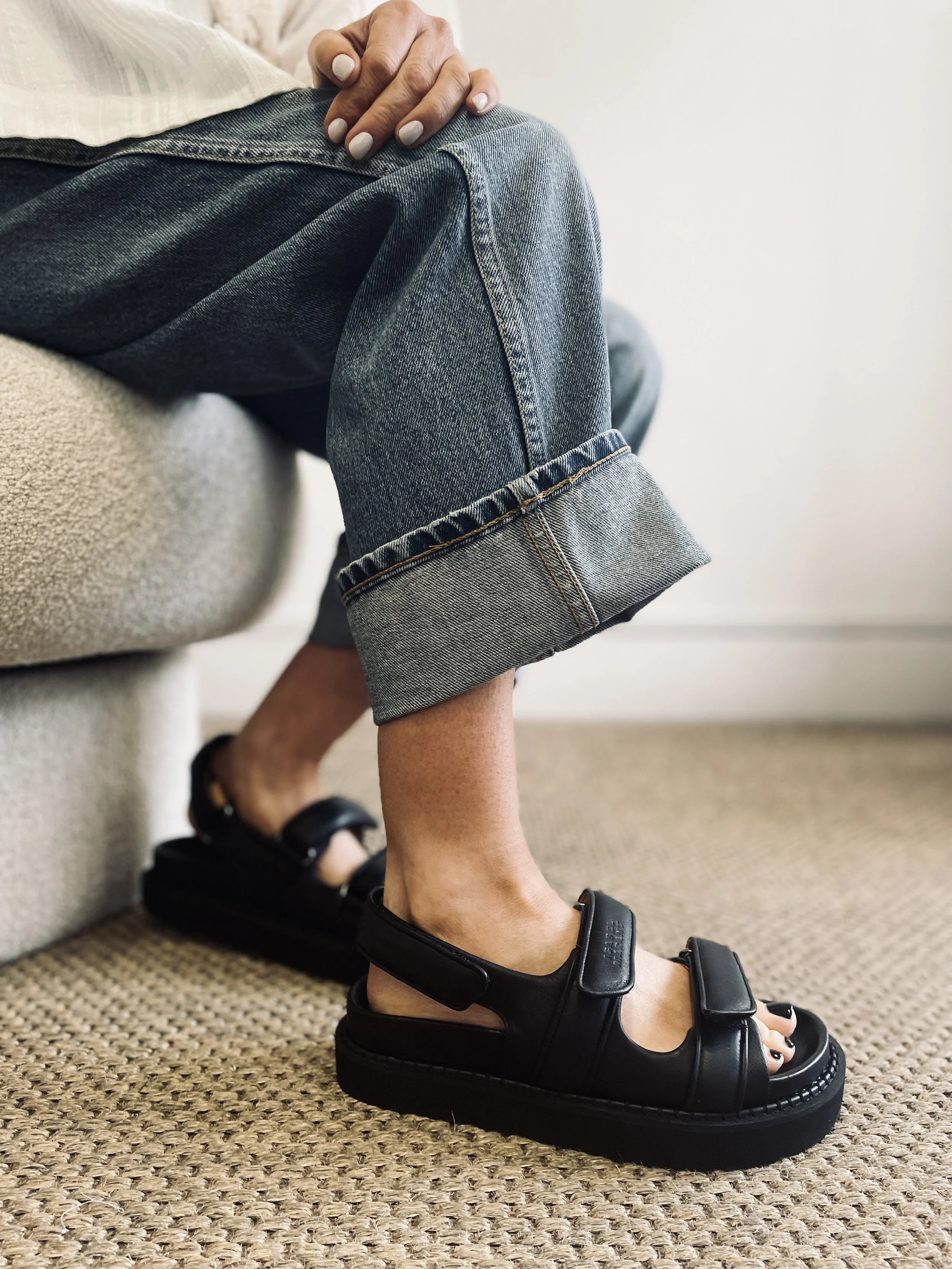 Person sitting on a couch, wearing rolled-up blue jeans and black platform sandals with Velcro straps, with their right foot on a woven rug.