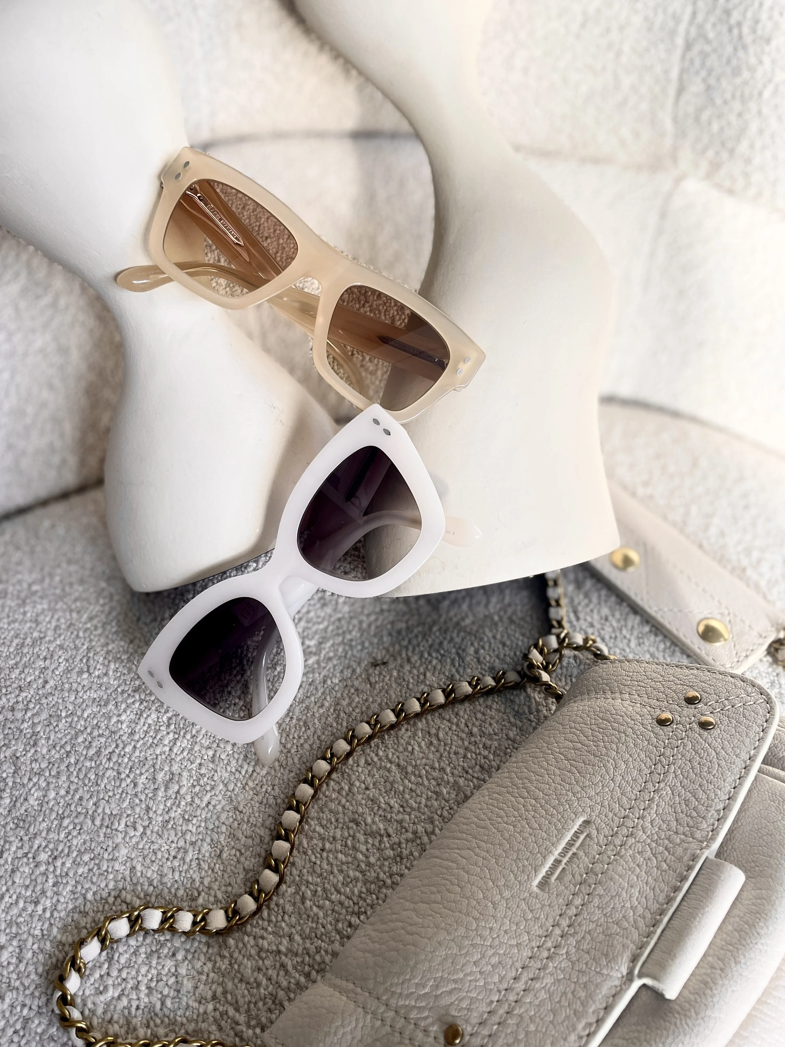 Two pairs of sunglasses, one beige with brown lenses and one white with black lenses, are placed on a beige hat. A beige textured clutch with gold chain detail and a white label is also visible.