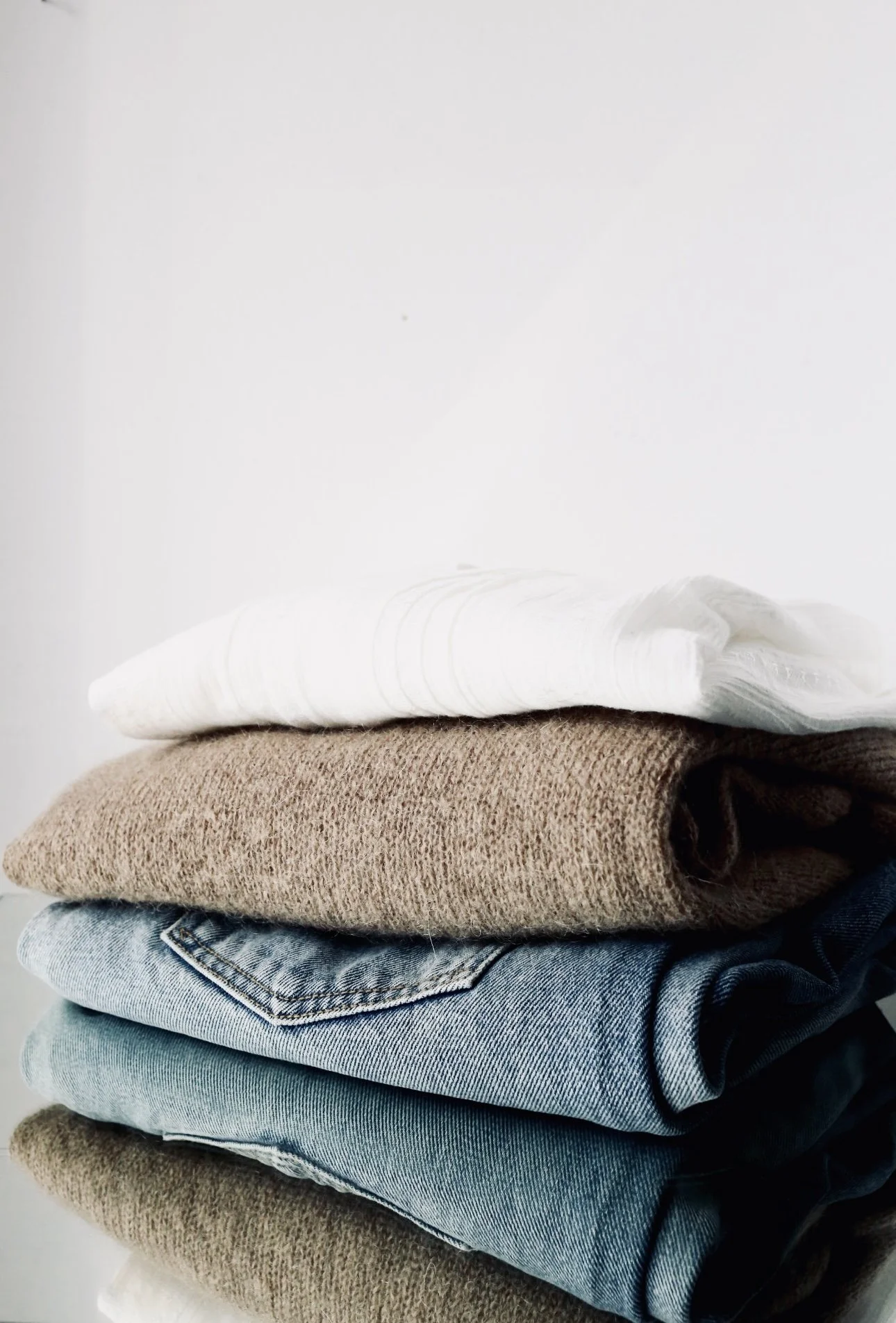 Stacked folded clothes including white, beige, blue jeans, and dark-colored pants against a white wall.