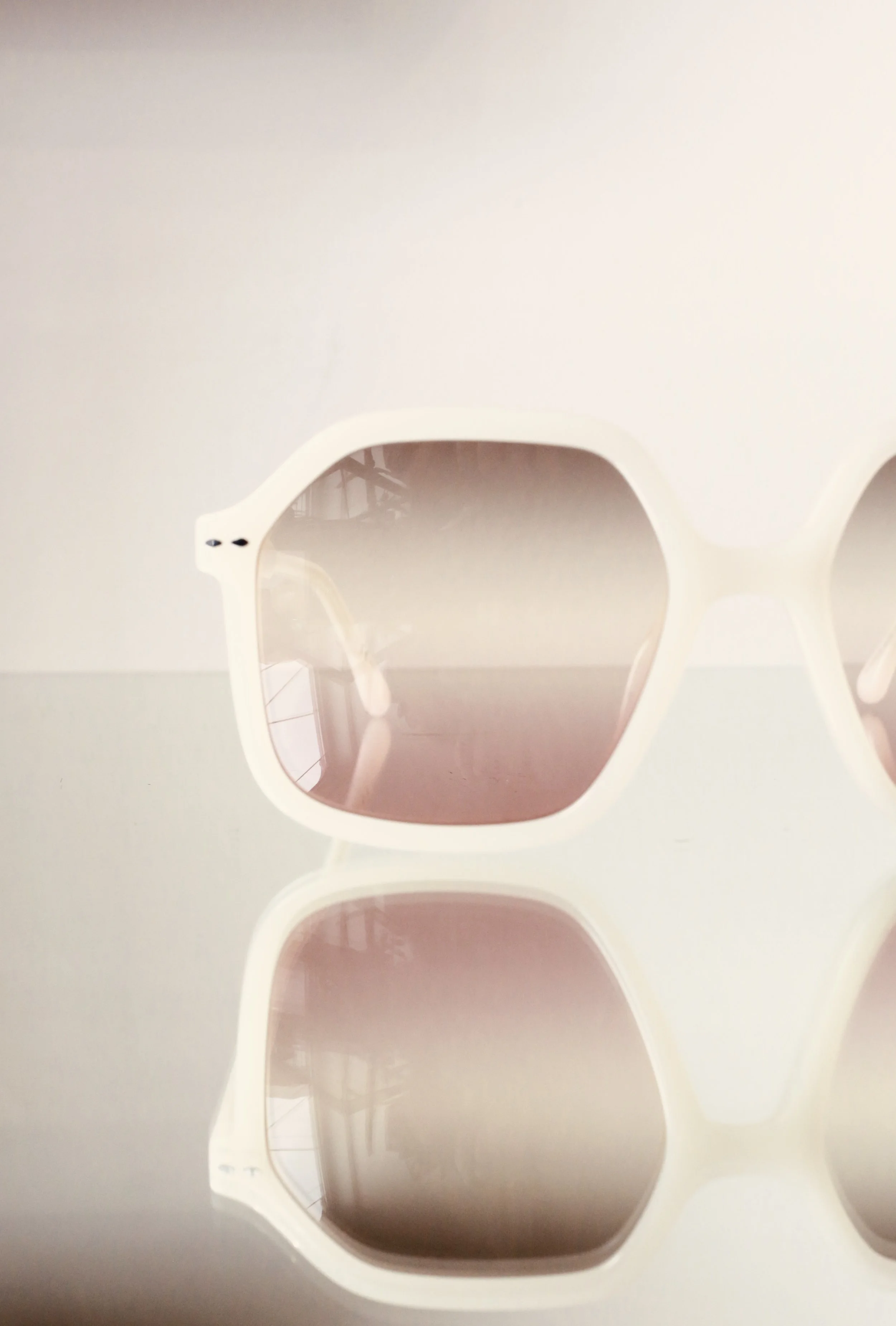 Close-up of white sunglasses with pink-tinted lenses on a reflective surface, showing a partial reflection of the surrounding environment.