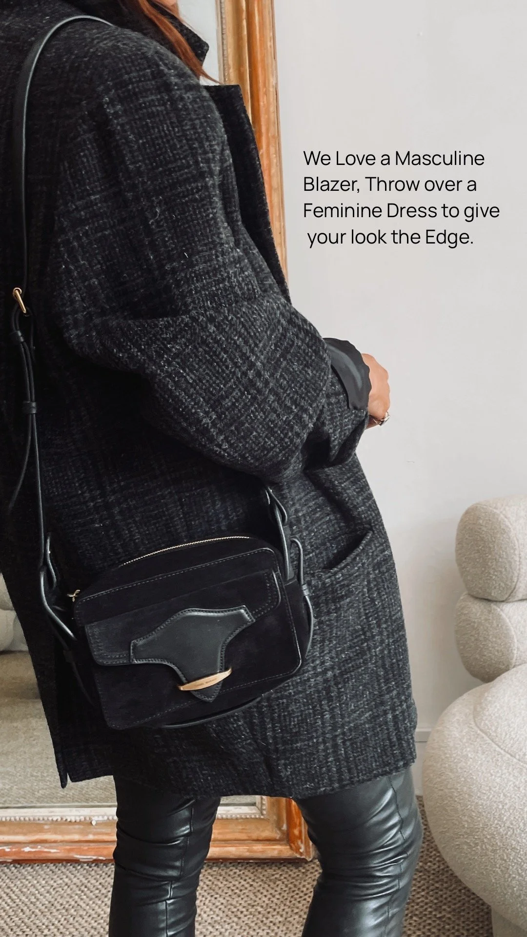 Person wearing a black blazer layered over a feminine dress, with a black crossbody bag, standing indoors near a beige sofa and a mirror, with a white wall in the background.