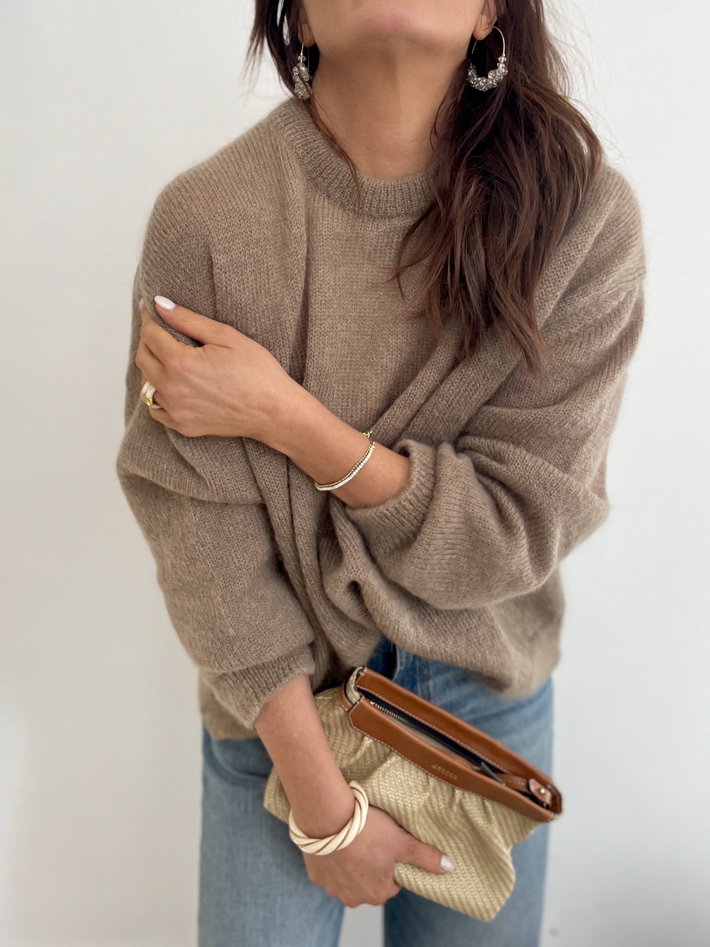A woman wearing a beige sweater, blue jeans, and jewelry including earrings, rings, a bracelet, and a bangle, holding a beige clutch and a brown wallet.