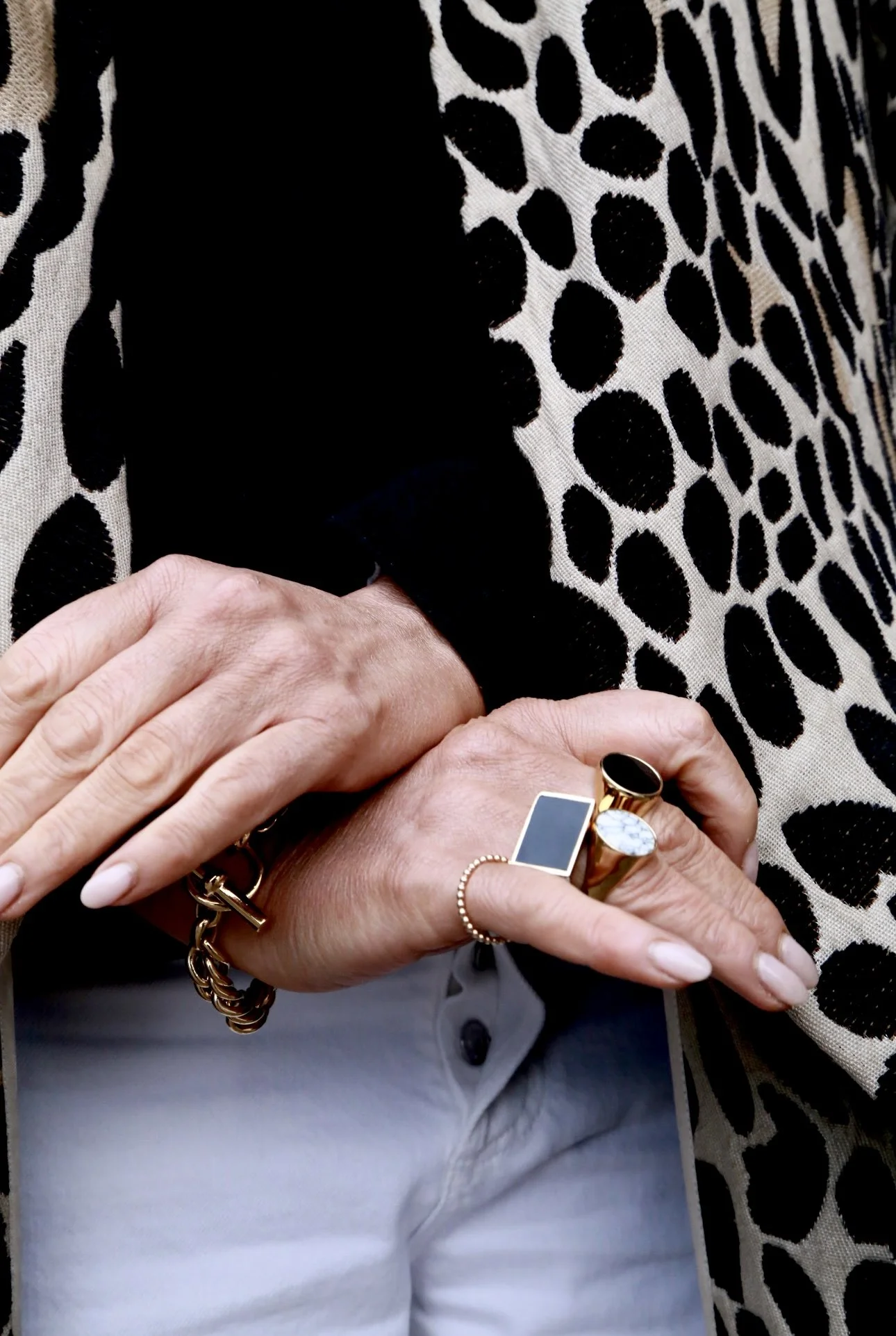Close-up of a person's hands with jewelry, including rings and a bracelet, against a leopard print fabric and part of a black and white top and light-colored pants.